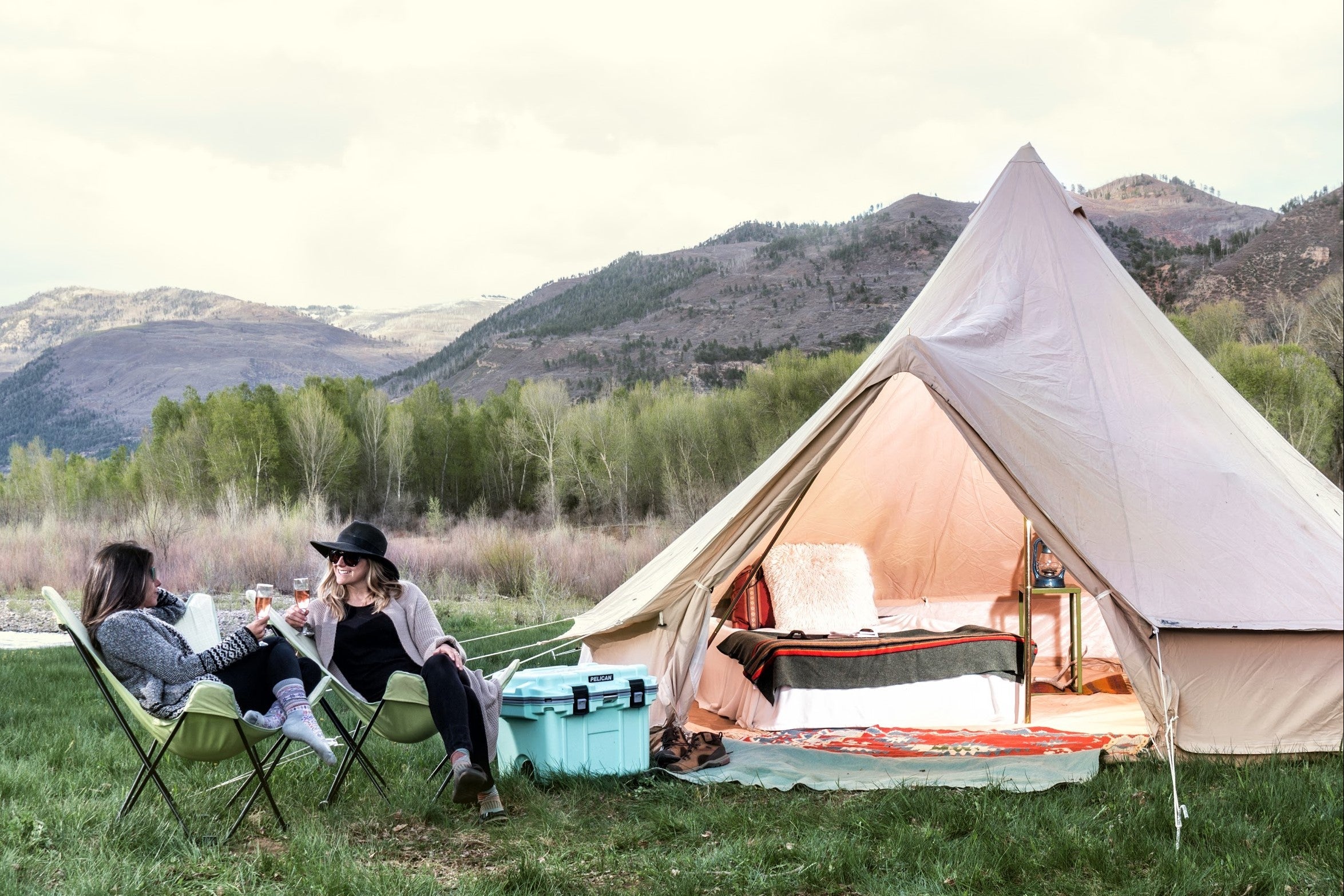 Two people sitting outside a tent in a scenic outdoor setting with mountains and trees.