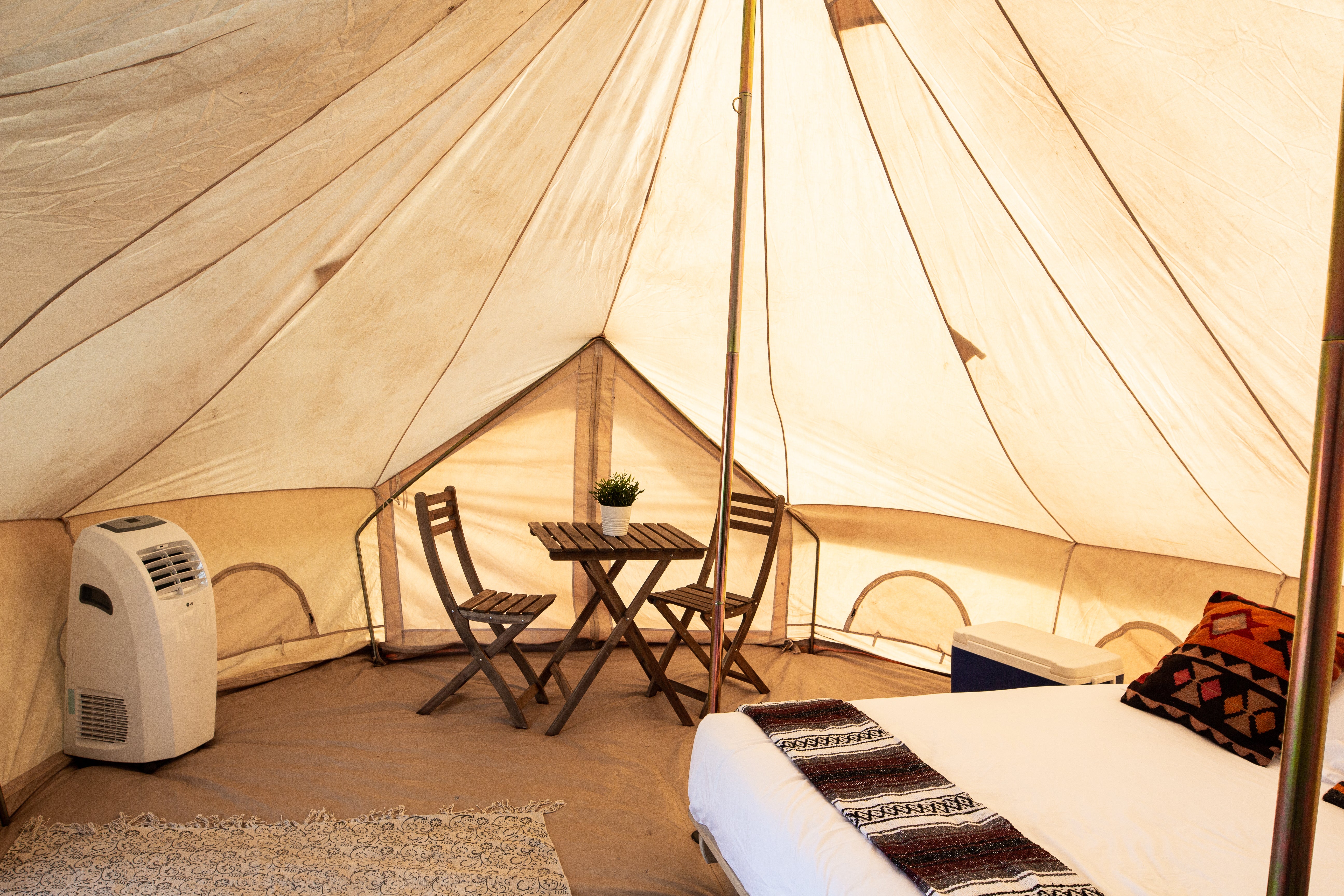 A cozy interior view of a large canvas glamping tent, featuring a small wooden dining table and chairs, a white bed with patterned textiles, a portable air conditioner, and warm ambient lighting.