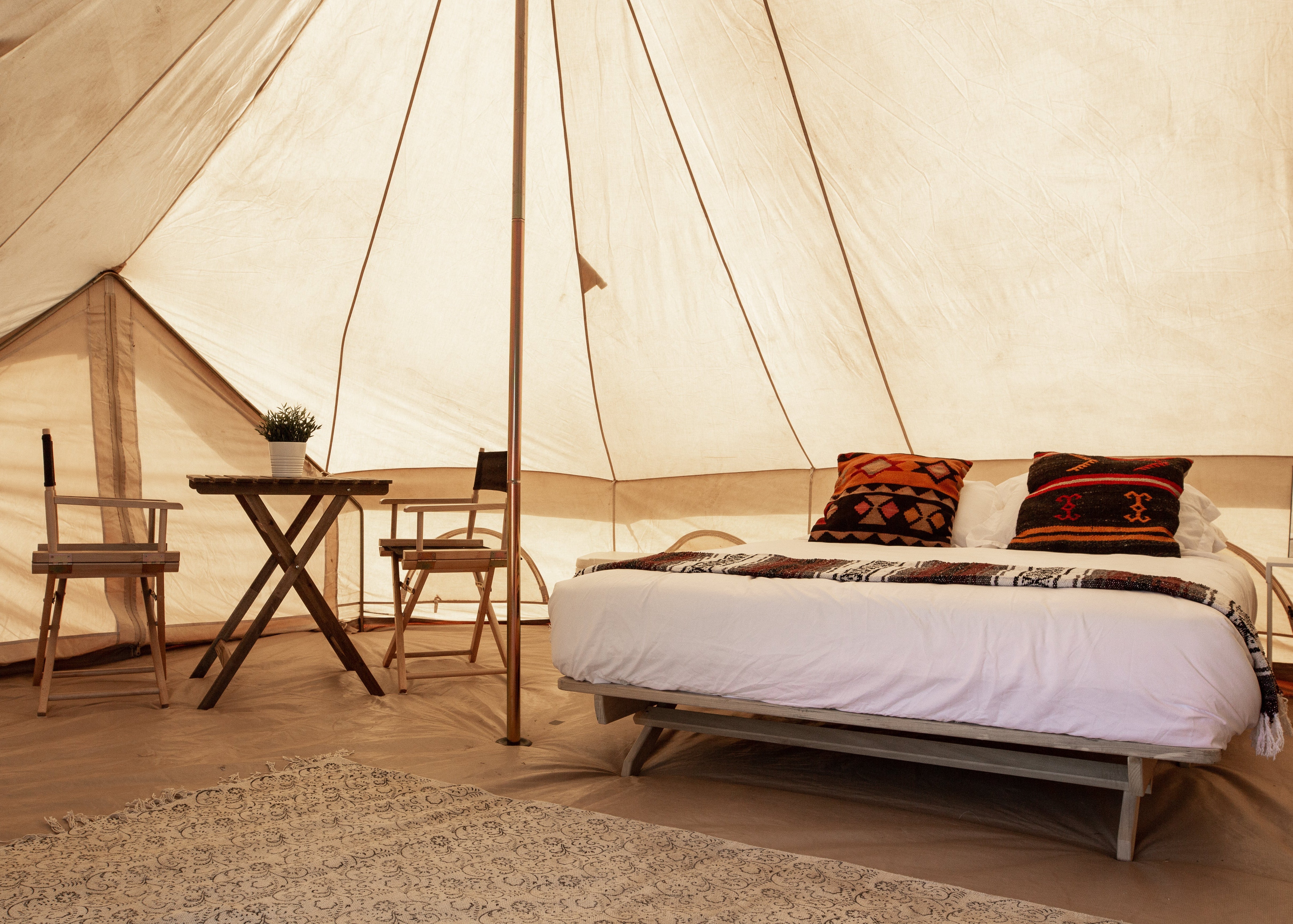 A luxury glamping tent bedroom featuring a large bed with white linens and patterned pillows, a small wooden folding table and chairs, and a textured rug inside a tall cream canvas tent.