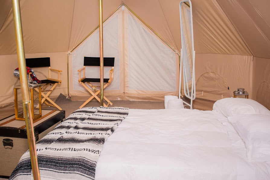 A luxury glamping tent bedroom featuring a large bed with white bedding and a black-and-white patterned blanket, two wooden director's chairs, and a full-length mirror inside a beige canvas tent.