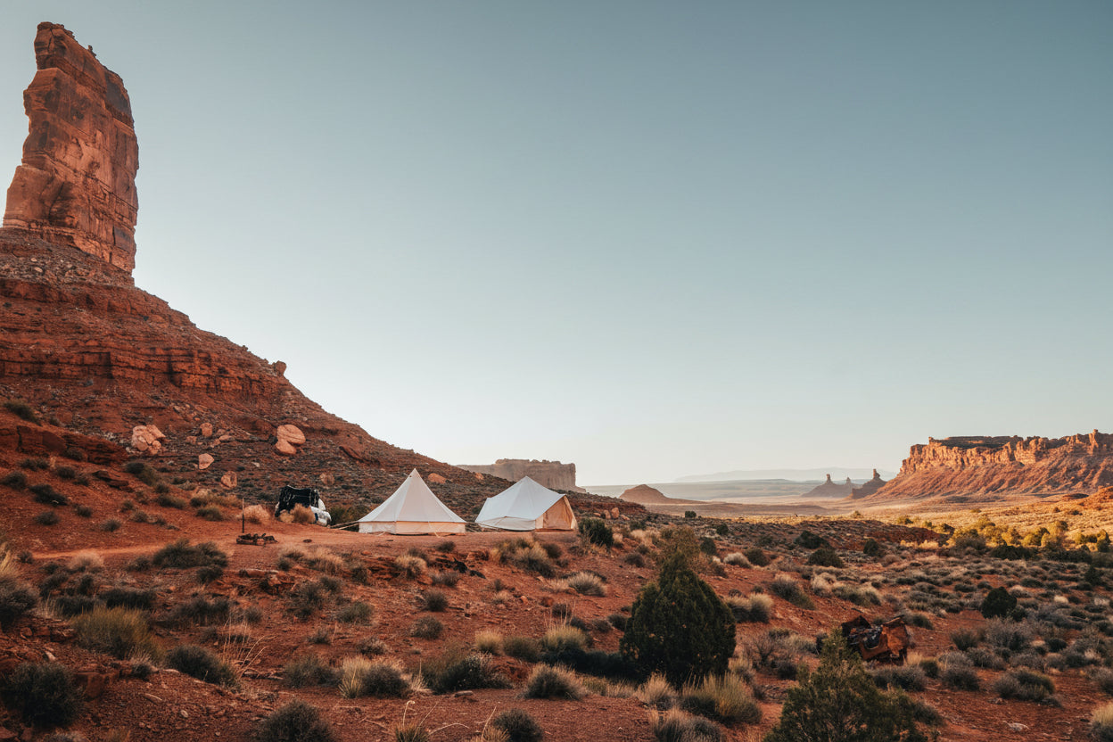 A desert landscape with red rock buttes and mesas under a pale blue sky, with two white tents pitched near the center and a small dark figure standing beside them.
