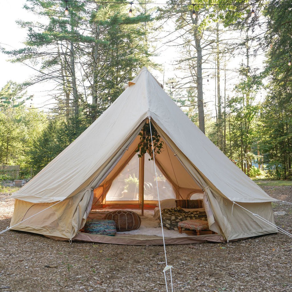 A large beige bell tent pitched outdoors in a wooded clearing, with trees behind it and a ground floor showing some bedding and decor inside.