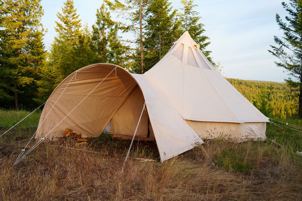A large canvas bell tent pitched outdoors in a grassy clearing with tall evergreen trees and a forested hillside in the background.
