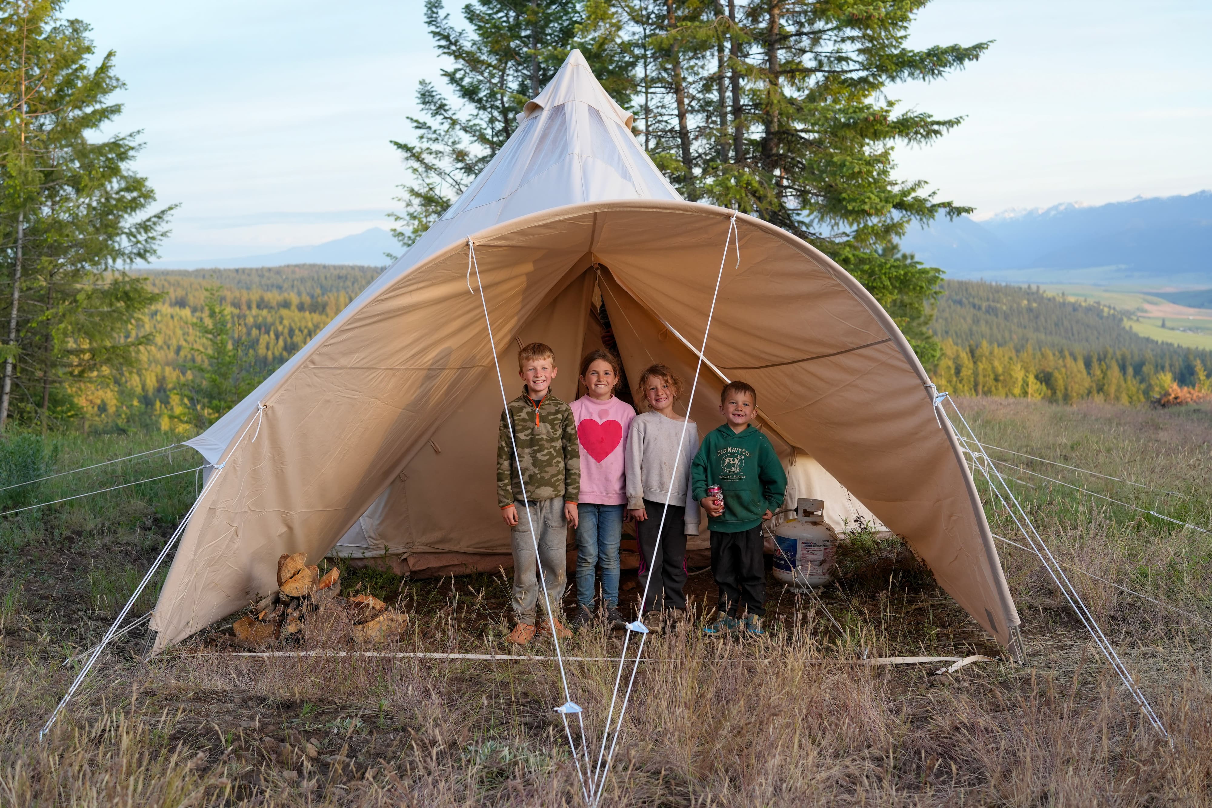 A large beige canvas bell tent pitched outdoors in a grassy mountain clearing, with four children standing inside the open front of the tent. Tall evergreen trees and distant mountains are visible in the background.