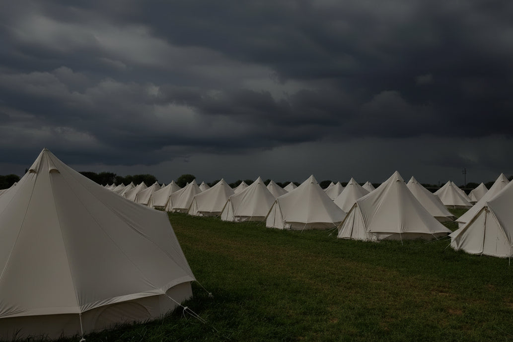 A row of large white bell tents pitched on a grassy field under a dramatic cloudy sky.