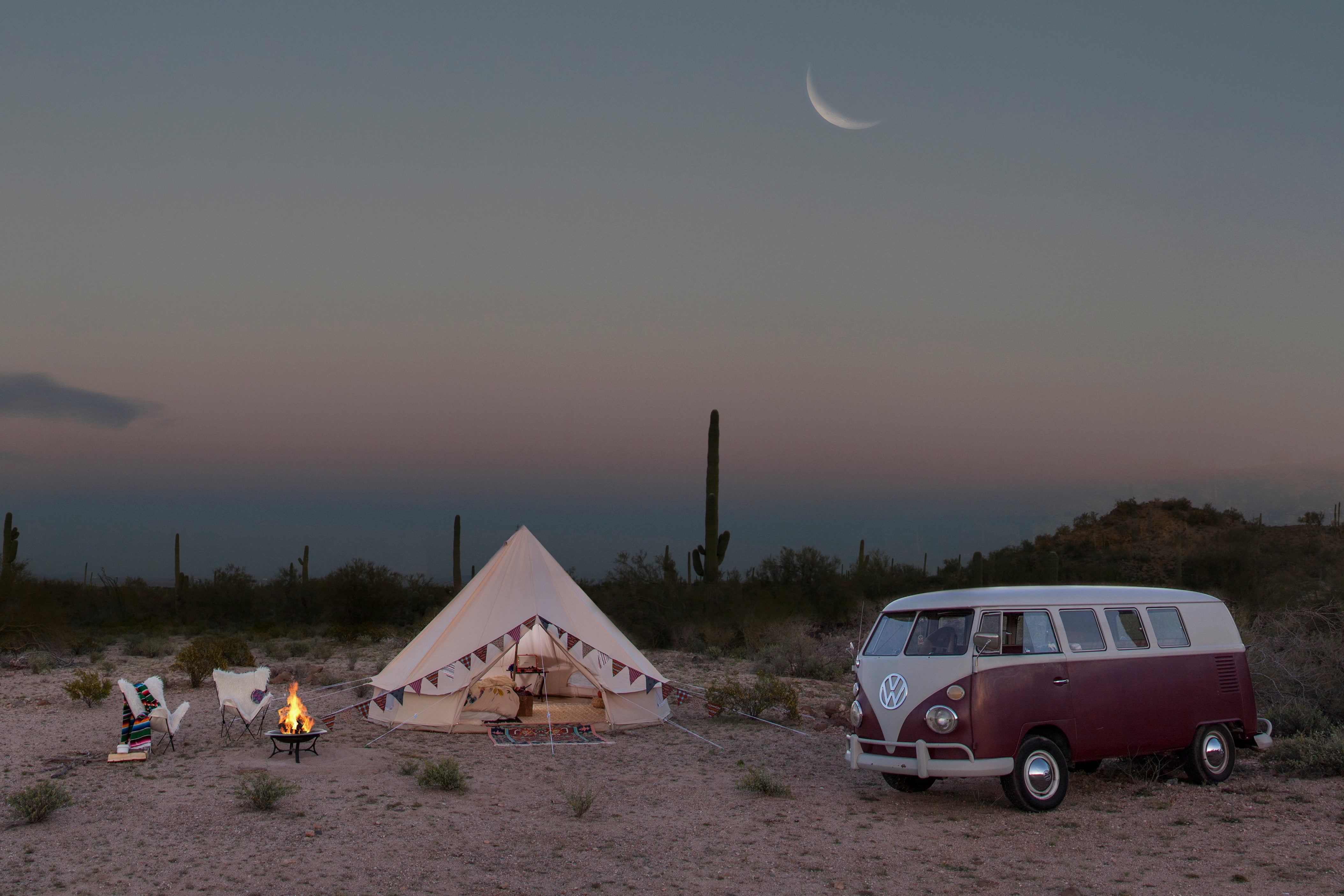 Desert scene with a tent, vintage van, and person by a campfire under a crescent moon.