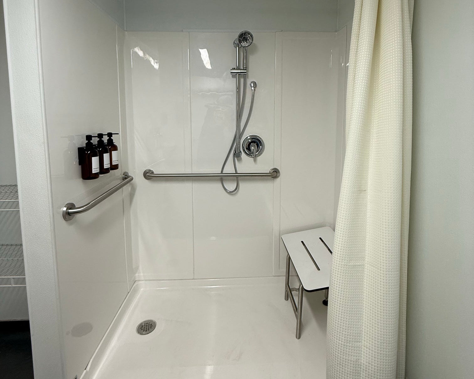 A modern white accessible shower stall with a built-in bench, grab bars, handheld shower, and a white textured shower curtain. The room has white walls and ceiling, with a dark floor.