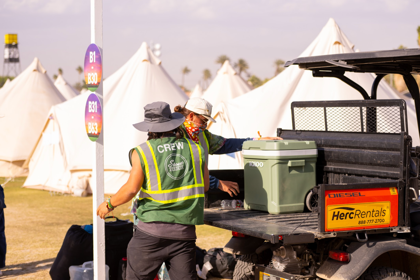 Two event crew members loading a cooler into a truck with tents and a clear sky in the background.