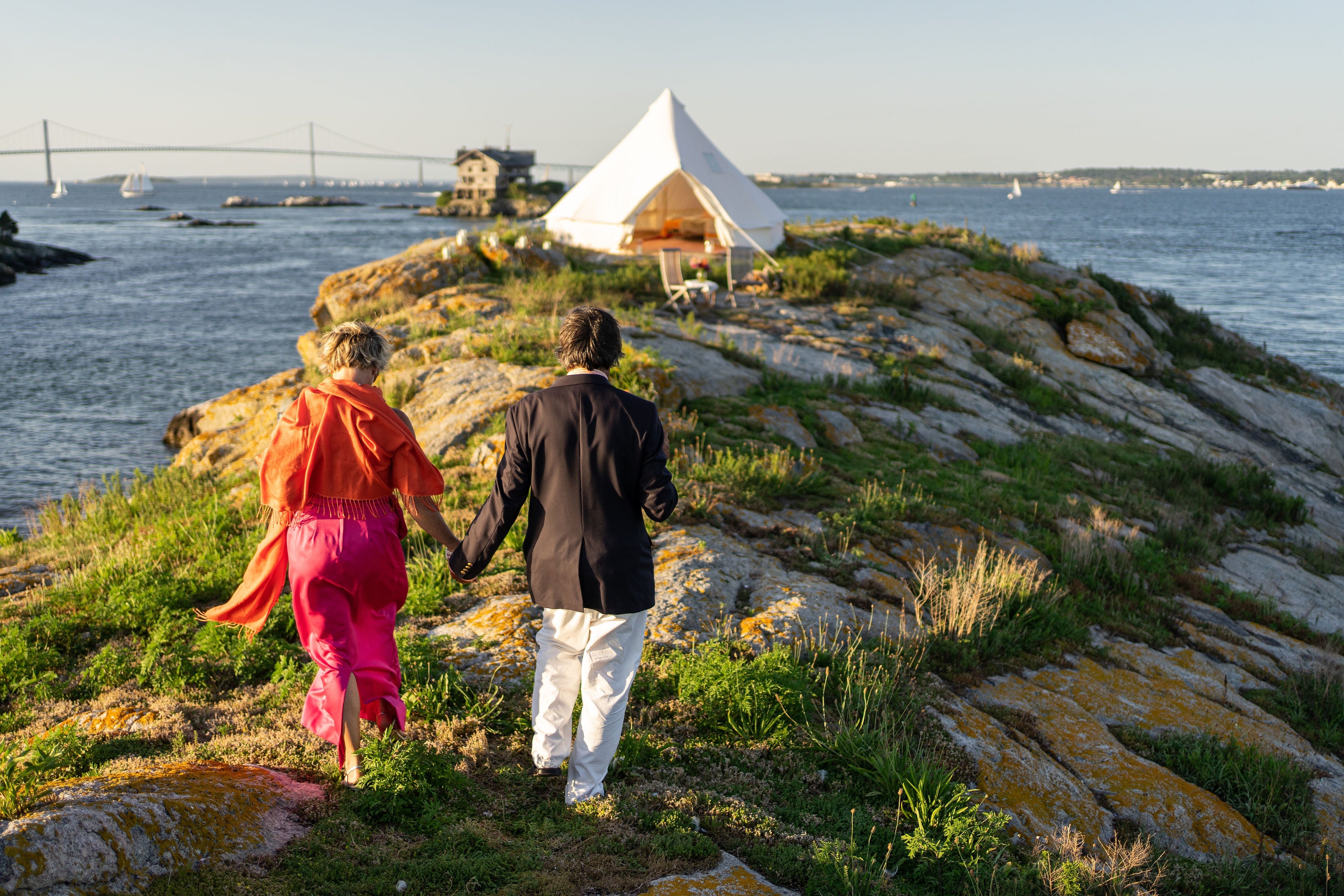 A couple holding hands walking toward a Stout Tent bell tent set up on a grassy, rocky cliffside overlooking the ocean.