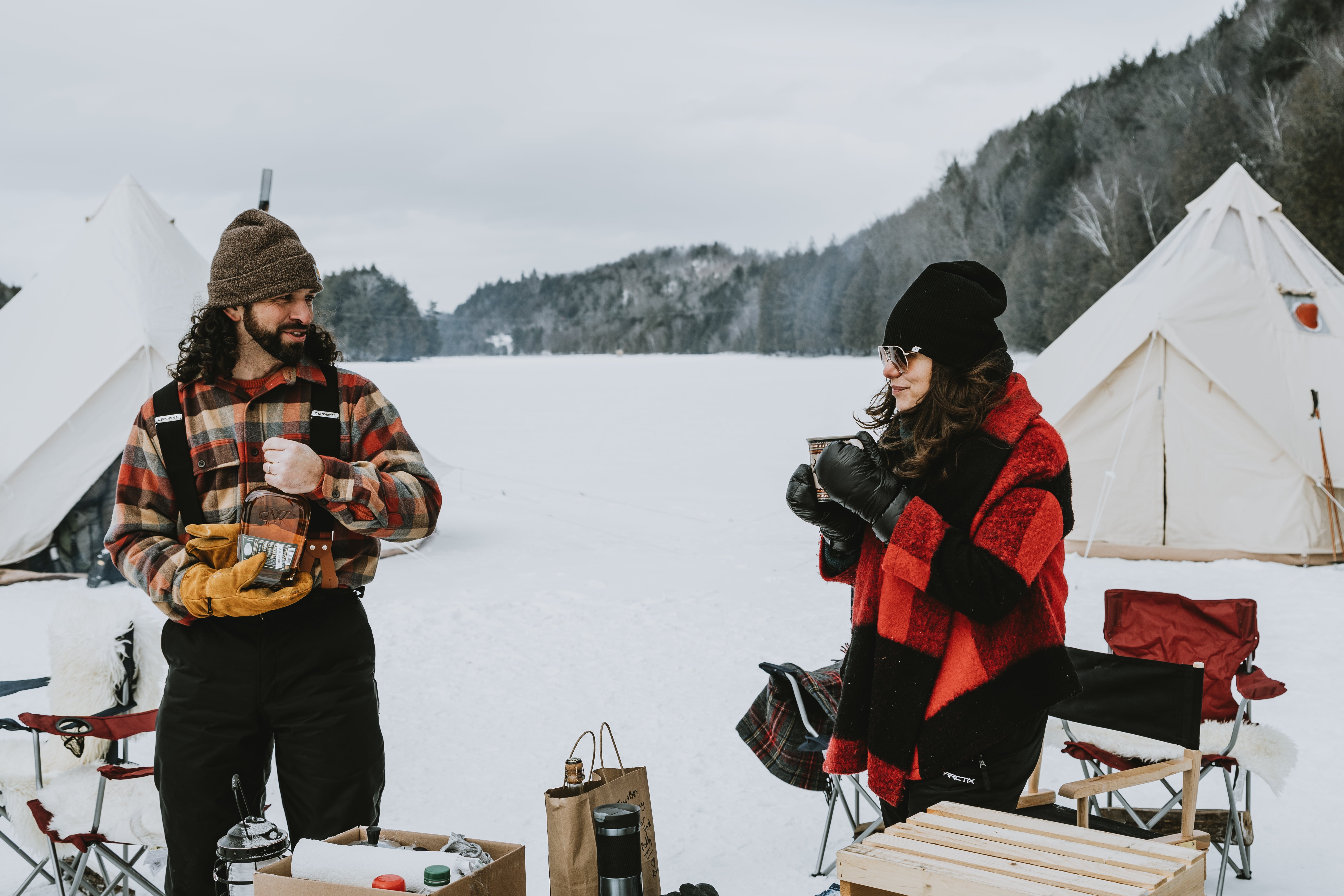 A couple in winter gear sitting outside a Stout Tent bell tent in a snowy landscape, holding mugs and enjoying the outdoors.
