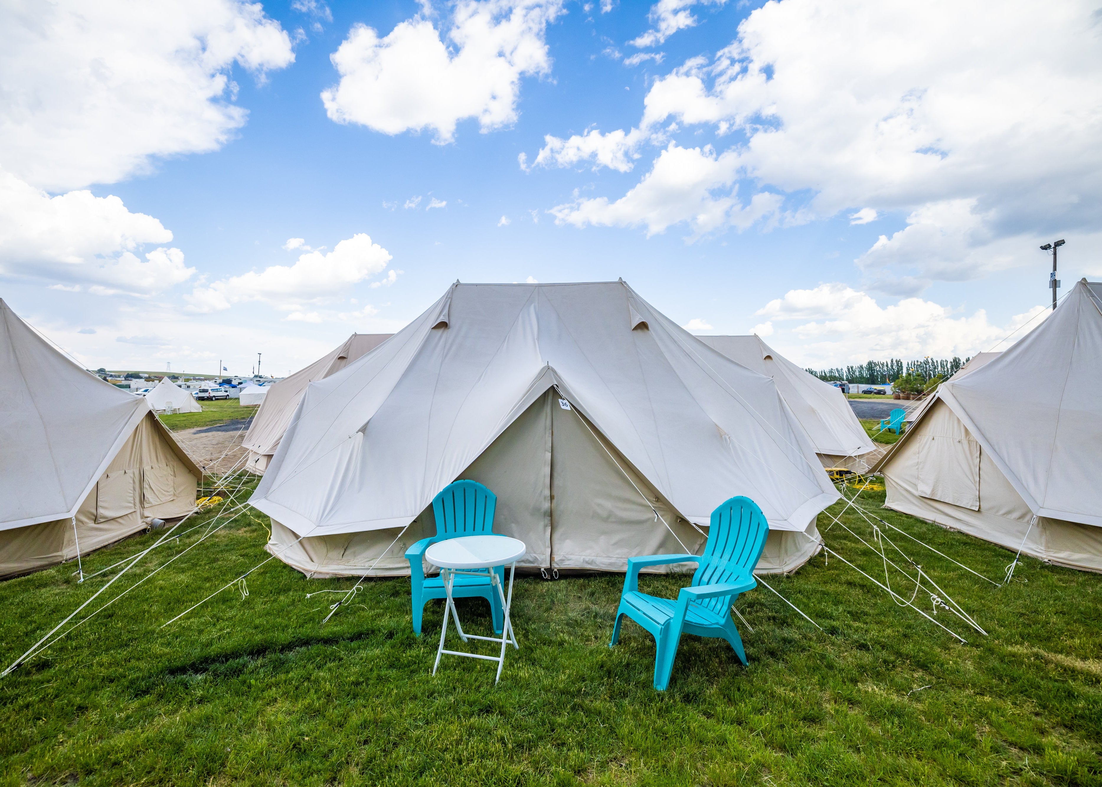 White tents with blue chairs and a table on a grassy field under a blue sky with clouds.