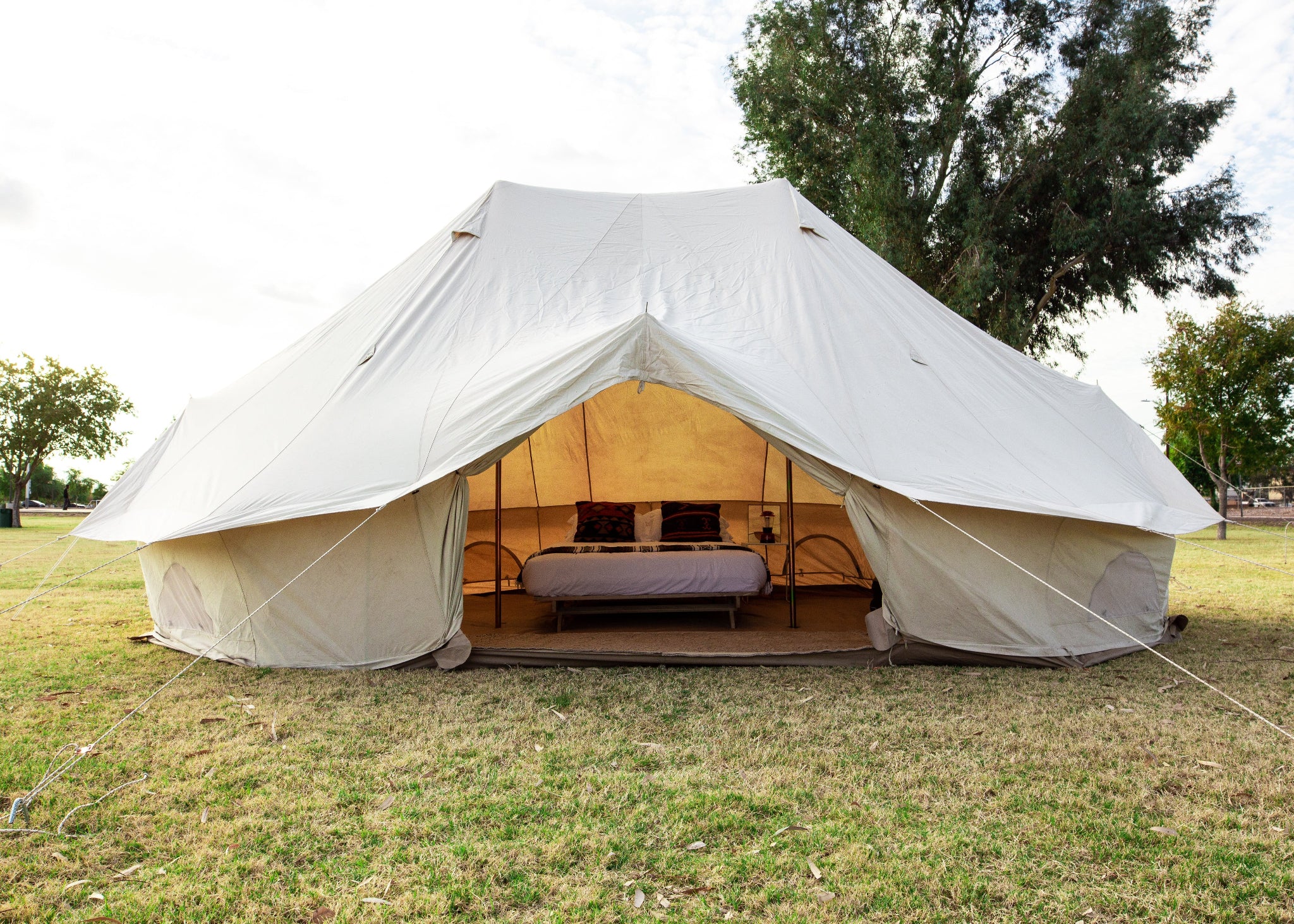 A large cream-colored canvas bell tent pitched on a grassy field with trees in the background. The tent is open, revealing a furnished interior with a bed and seating.