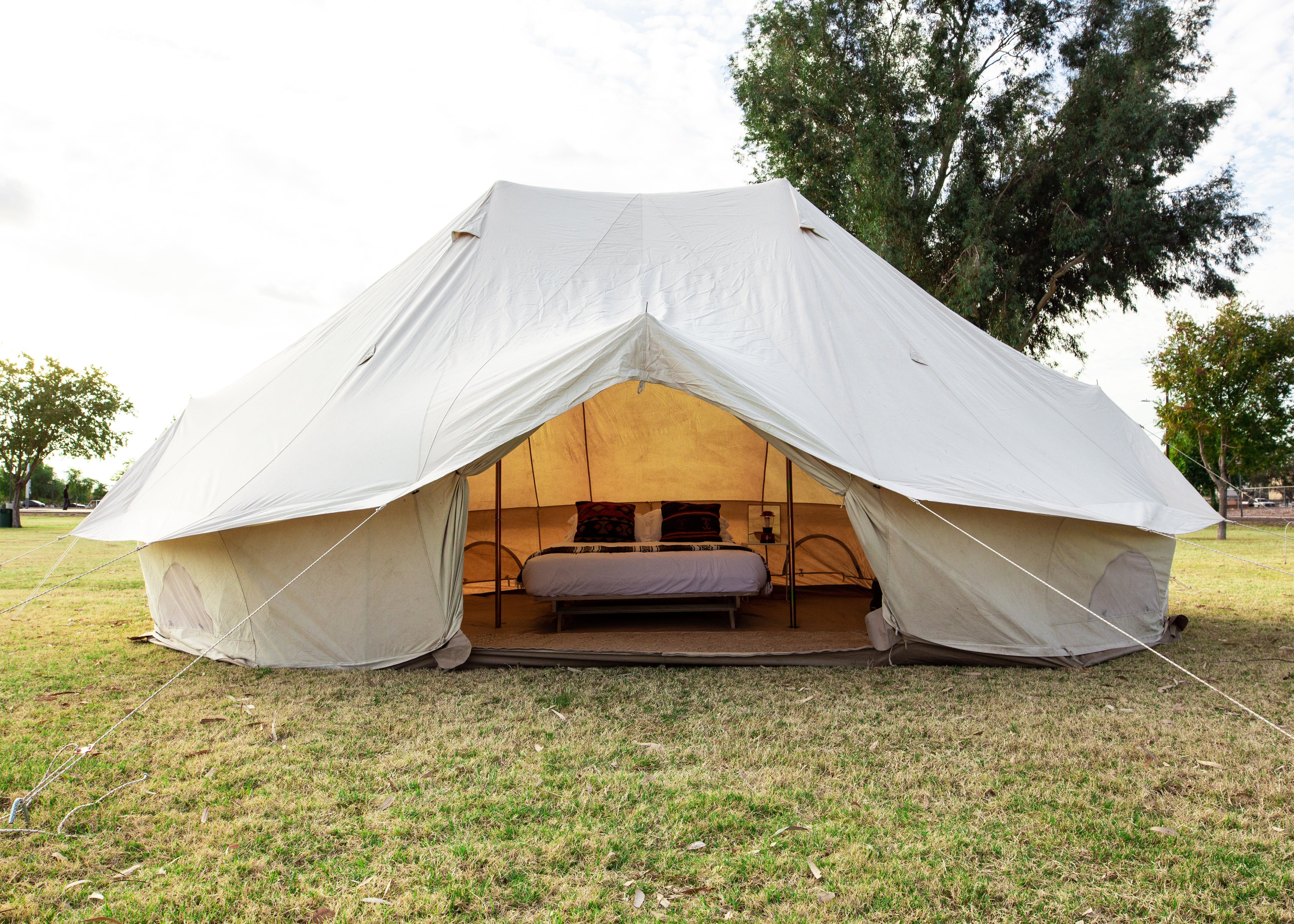 A large cream-colored canvas bell tent pitched on a grassy field with trees in the background. The tent is open, revealing a furnished interior with a bed and seating.