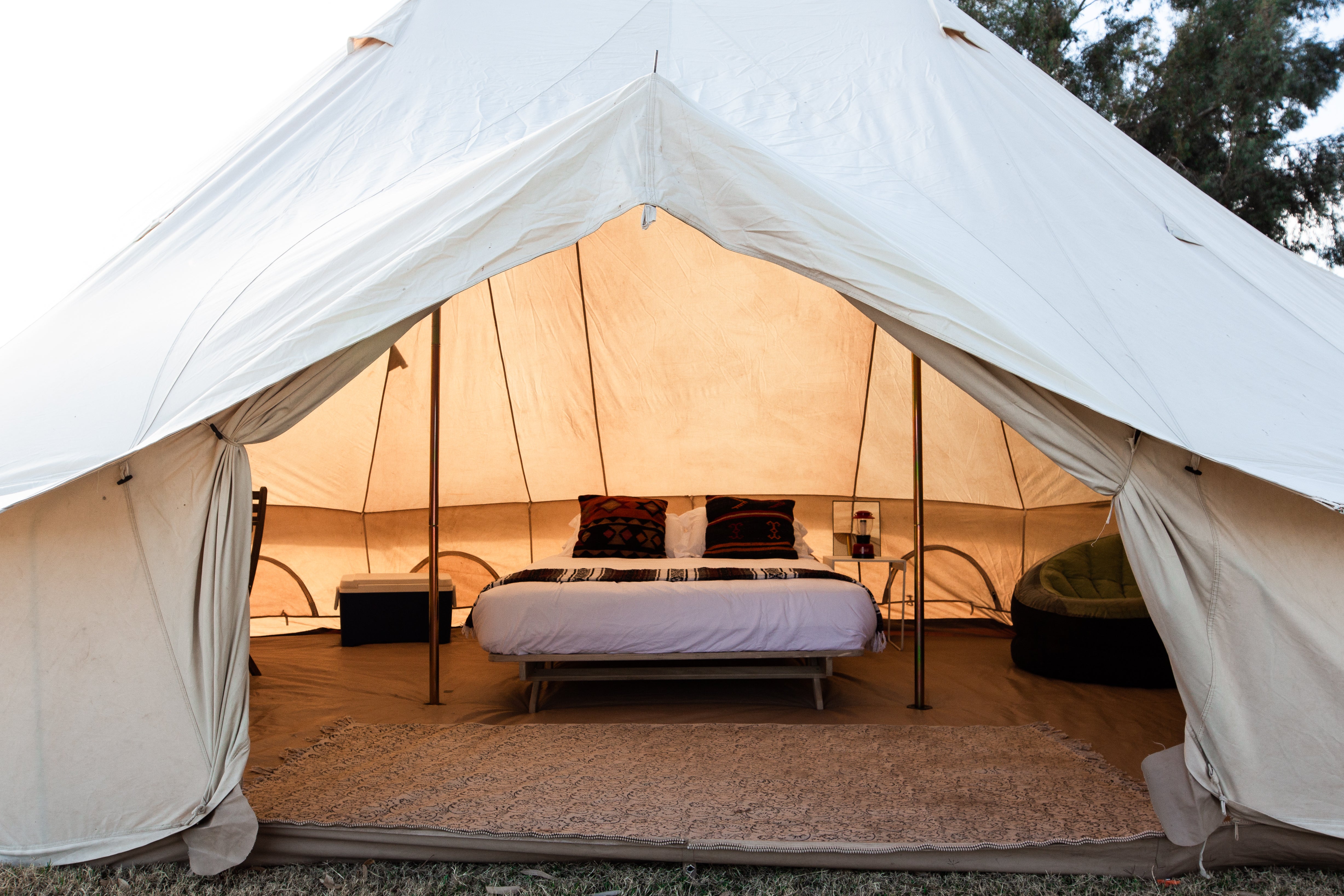 A large cream canvas bell tent with an open front, showcasing a fully furnished interior with a bed, side tables, and a green lounge chair on a carpeted floor.