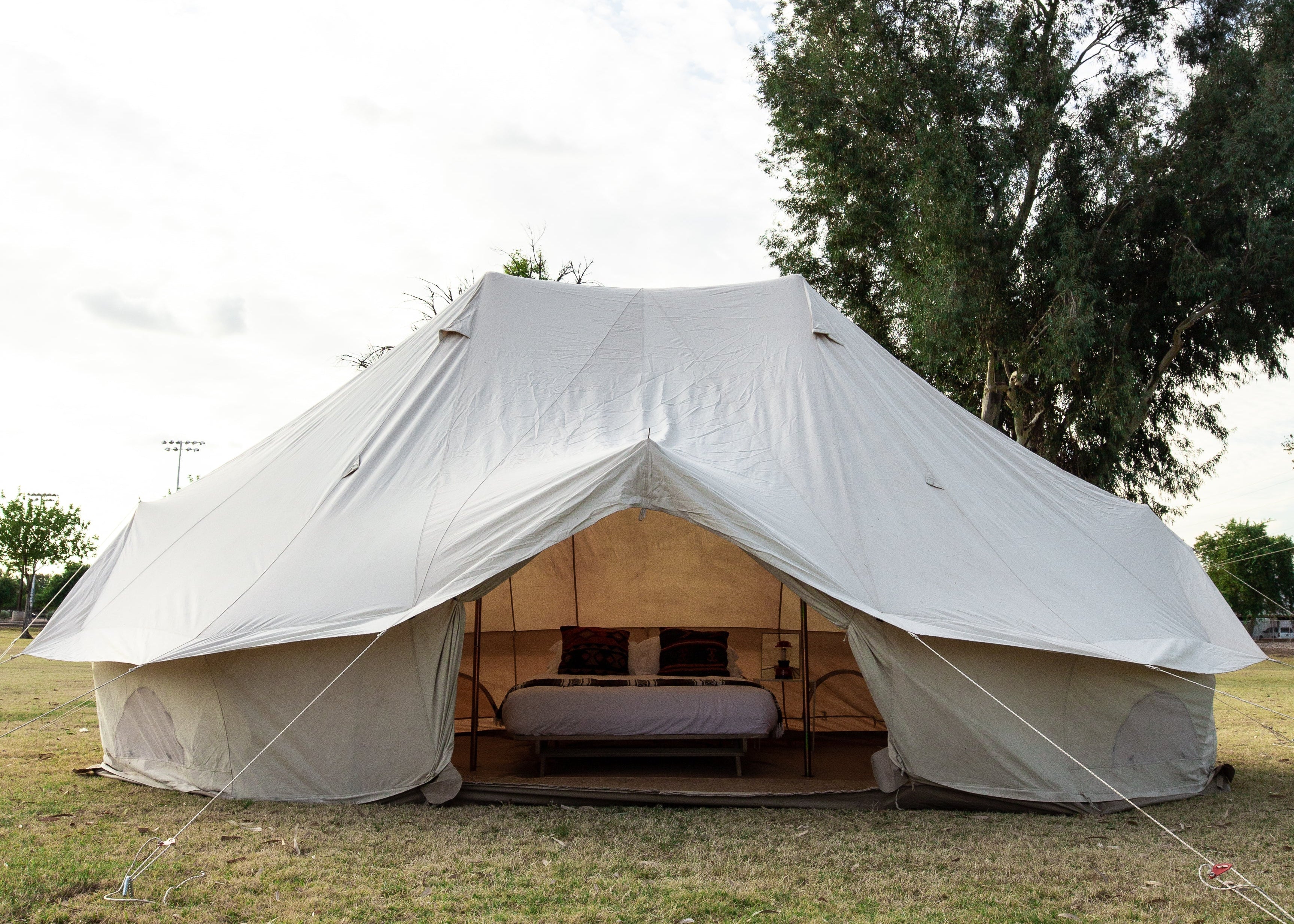A large canvas glamping tent with a wooden bed frame inside, pitched on a grassy field under a cloudy blue sky with trees in the background.