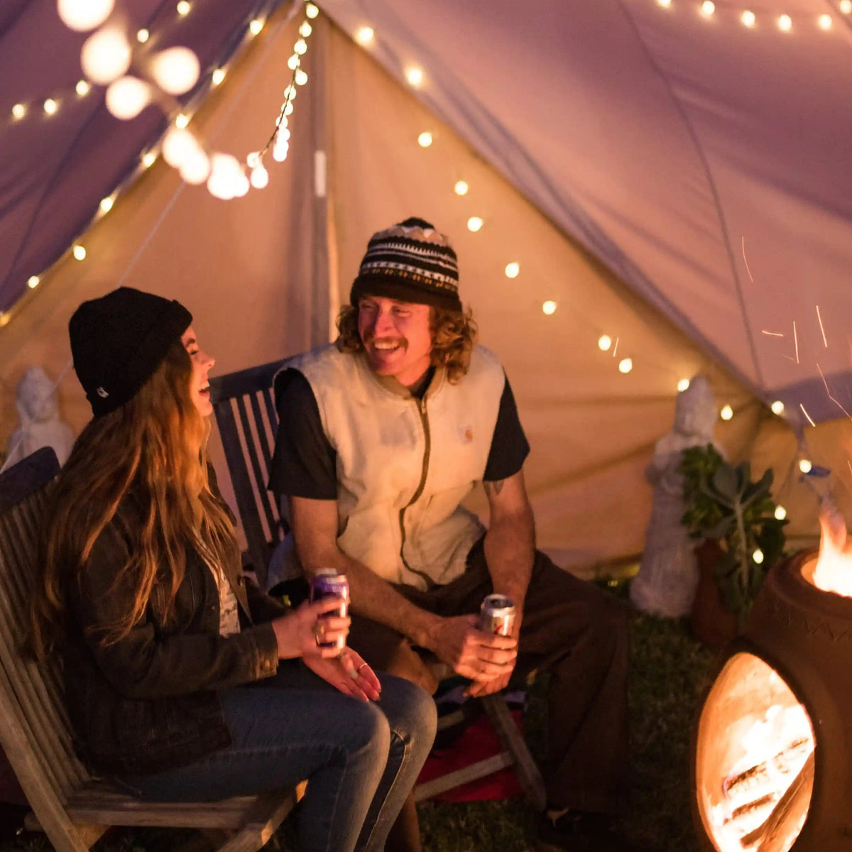 Two people sitting under a lit tent with string lights, near a fire pit.