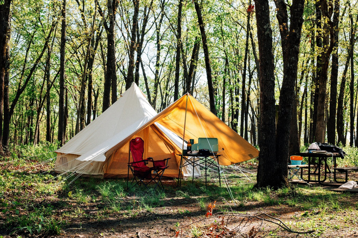 A tent with an awning set up in a forest with chairs and tables around.