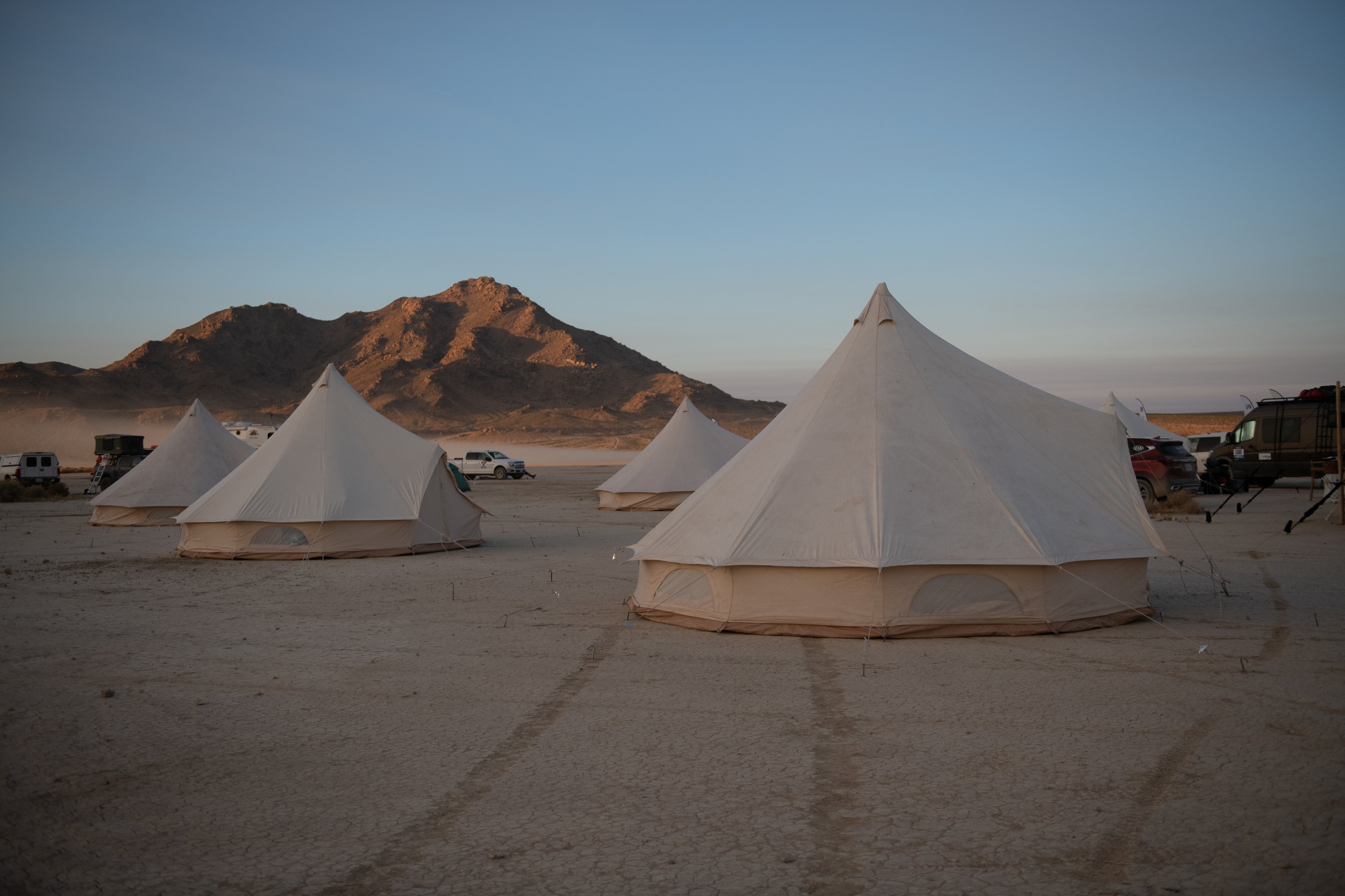 A large cream-colored canvas bell tent stands on a flat desert ground, with several smaller similar tents and vehicles arranged in the background near a mountain under a clear blue sky.
