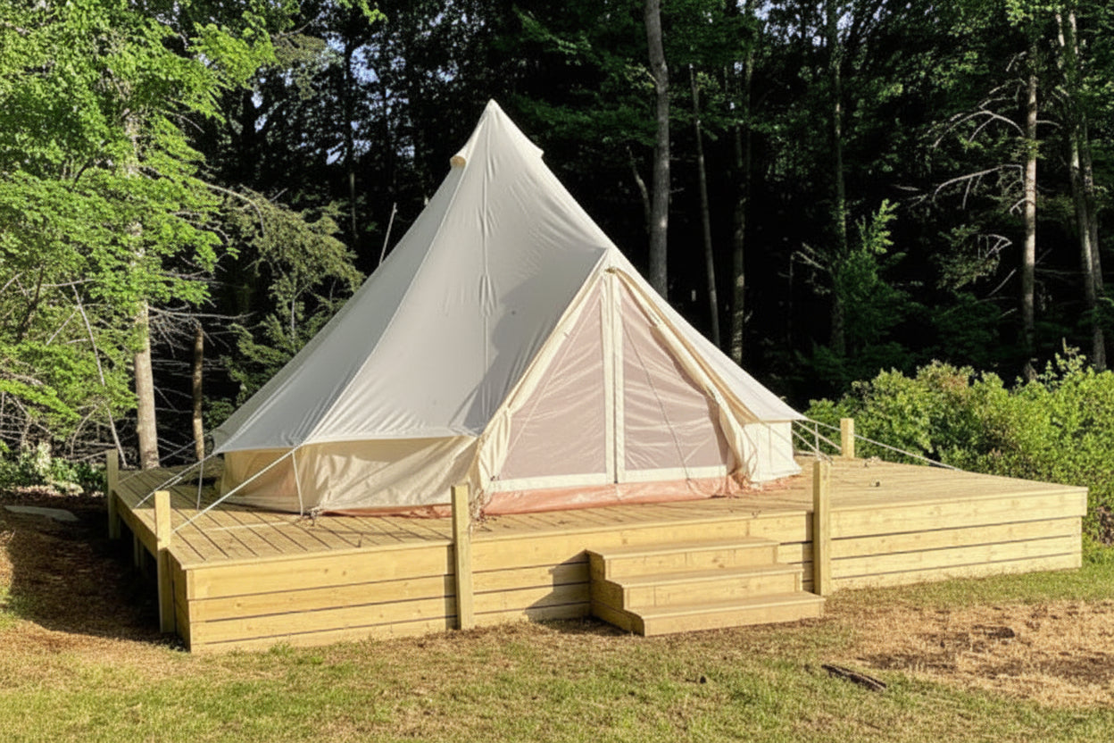 A large beige bell tent with a wooden platform and steps, set in a grassy clearing surrounded by a dense forest.