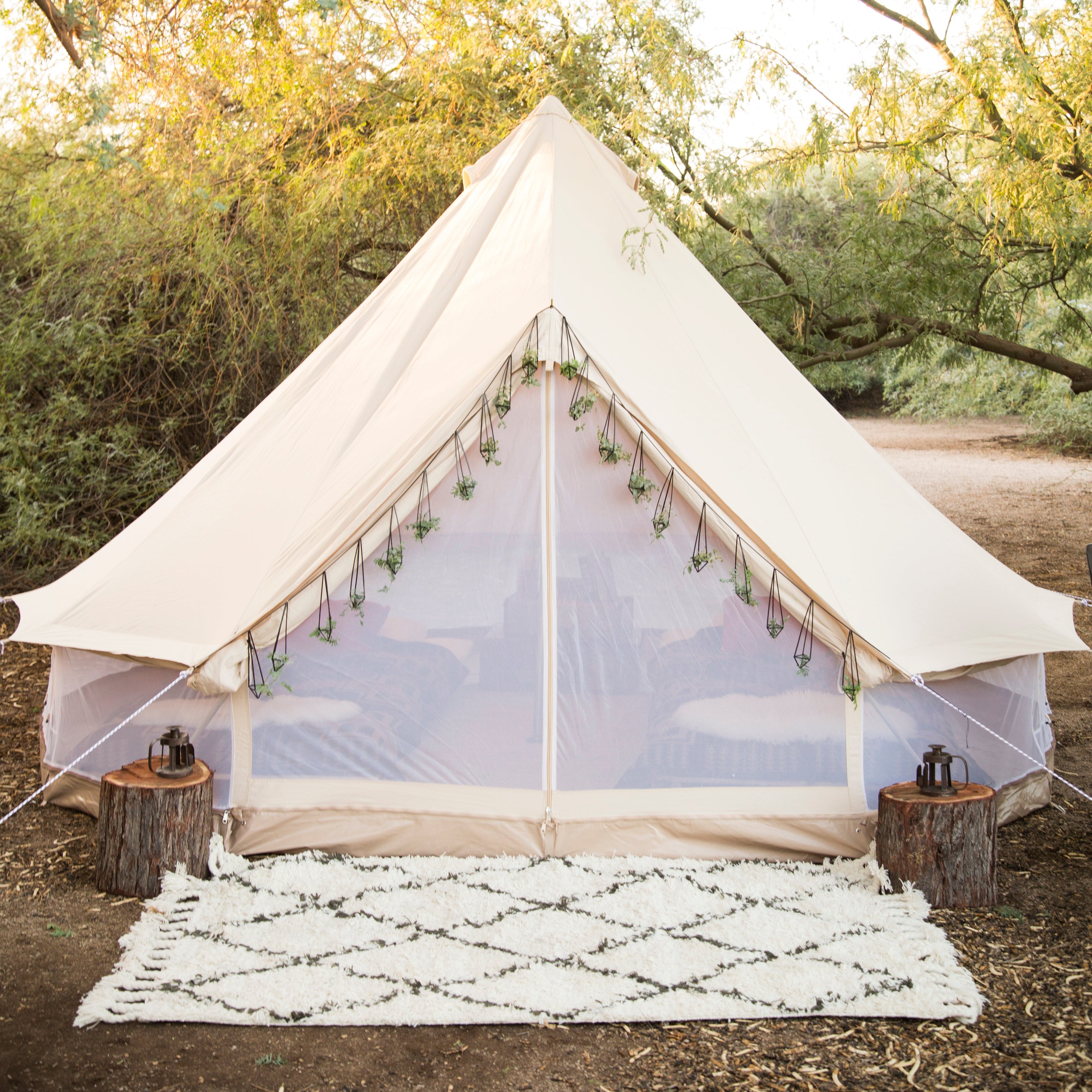 White bell tent with transparent sides in a natural setting with trees and a rug.