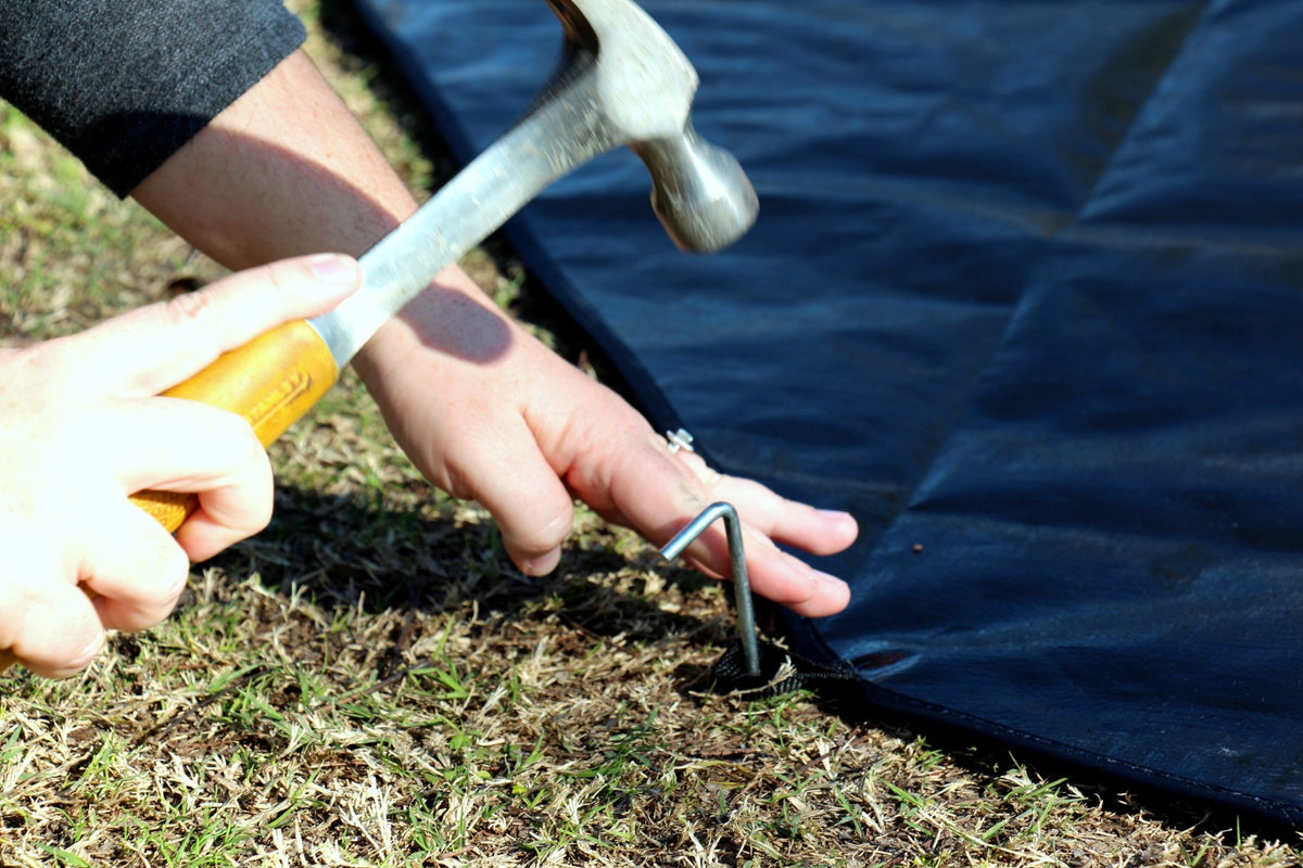 Person using a hammer to secure a metal stake into the ground next to a blue tarp.
