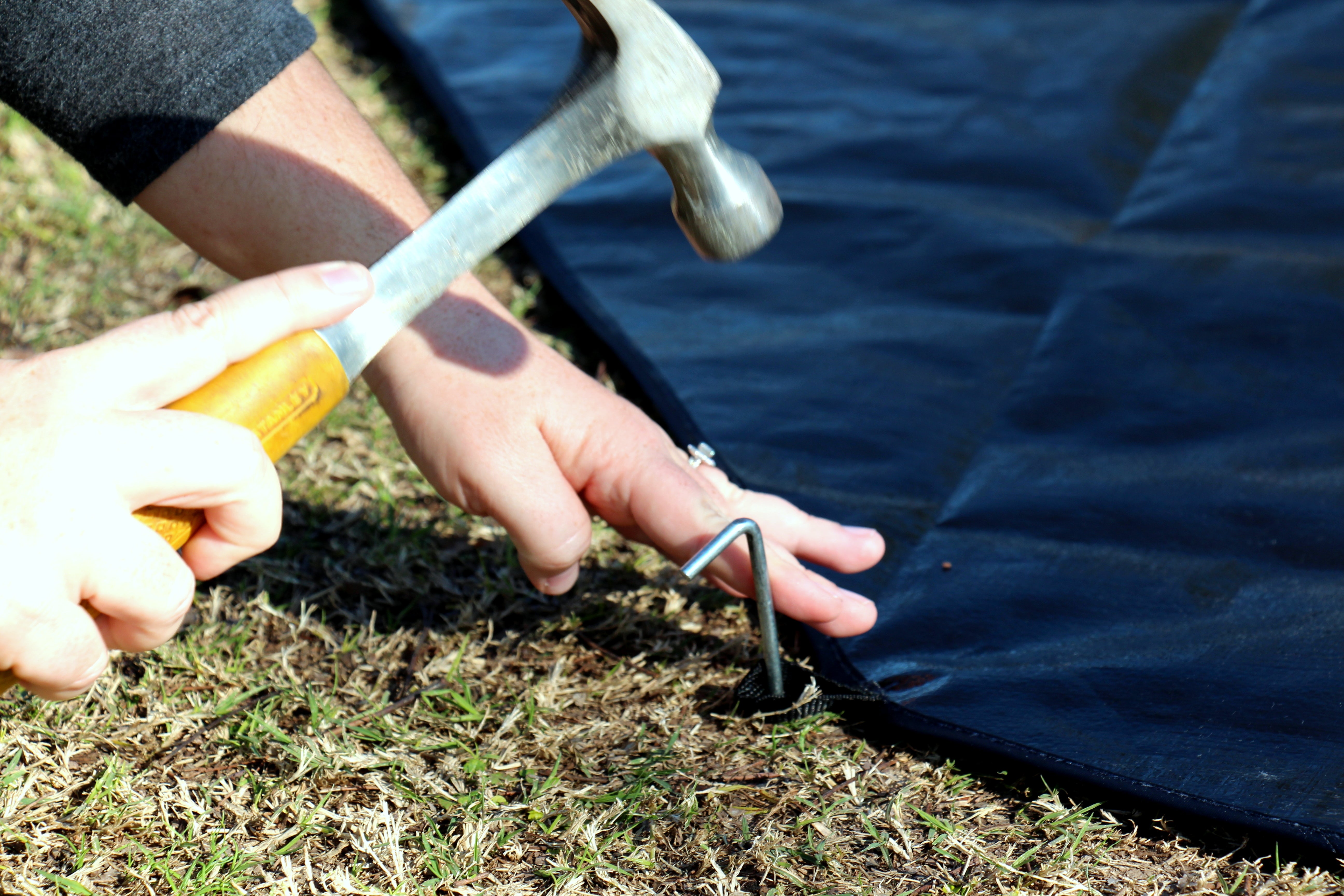 Person using a hammer to secure a metal stake into the ground next to a blue tarp.
