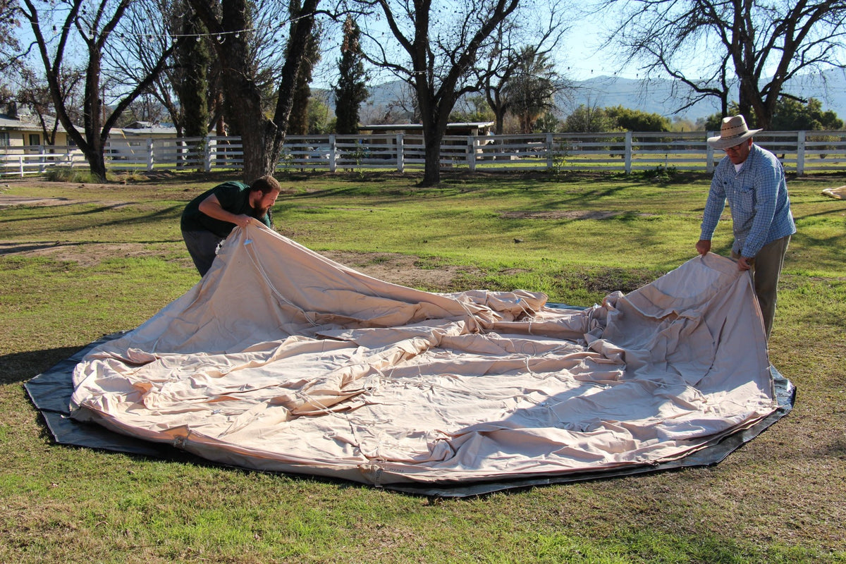 Two people working on a large beige tarp in an open field with trees and a fence in the background.