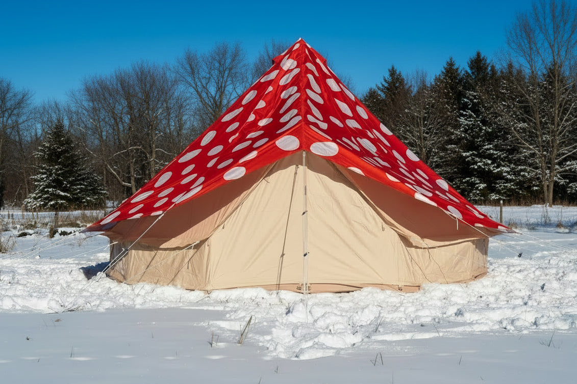 A large bell tent pitched outdoors in a snowy field under a blue sky. The tent has a red fly with white polka dots on the top and a light beige body.