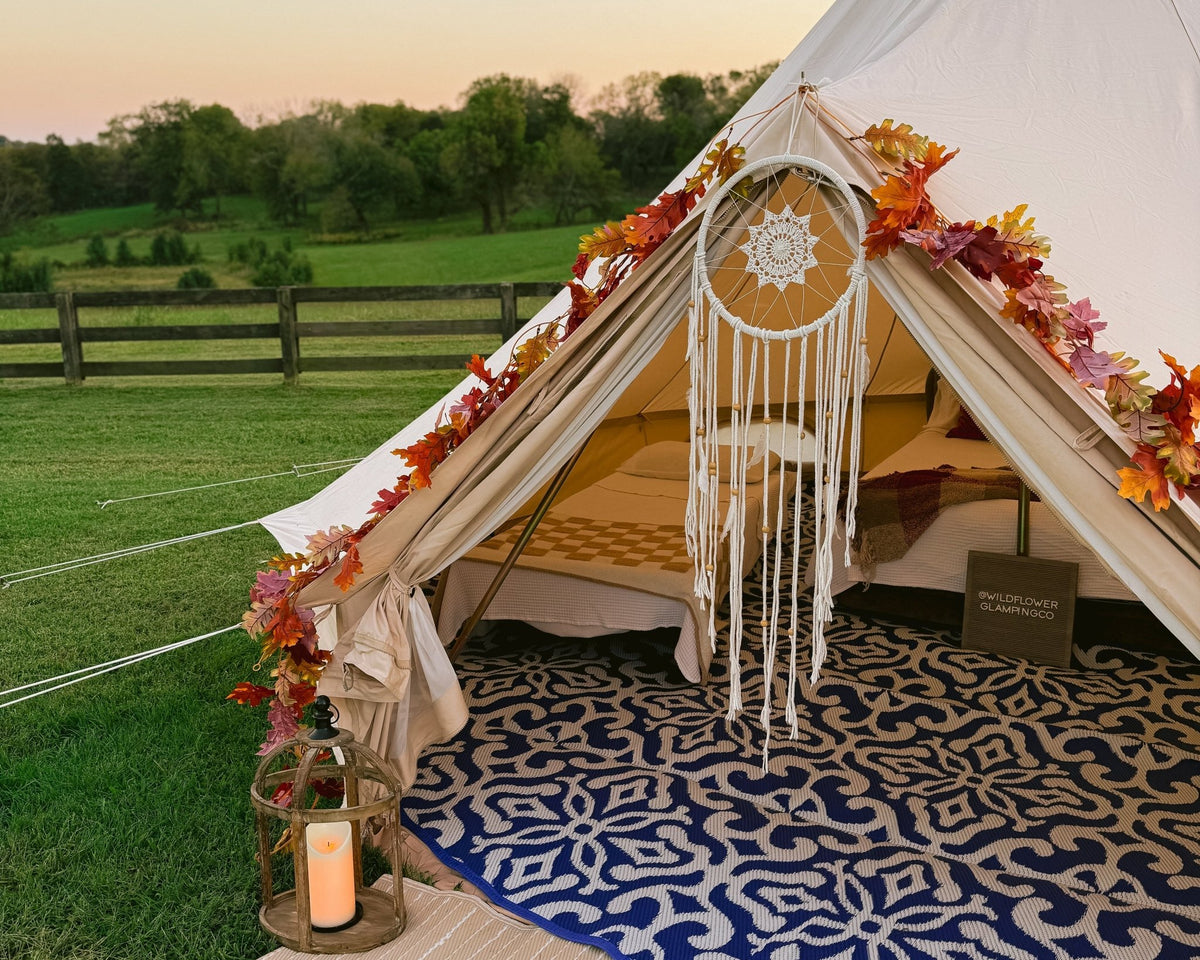 Decorated tent with fall leaves, a rug, and lantern on a grassy field with a wooden fence in the background.