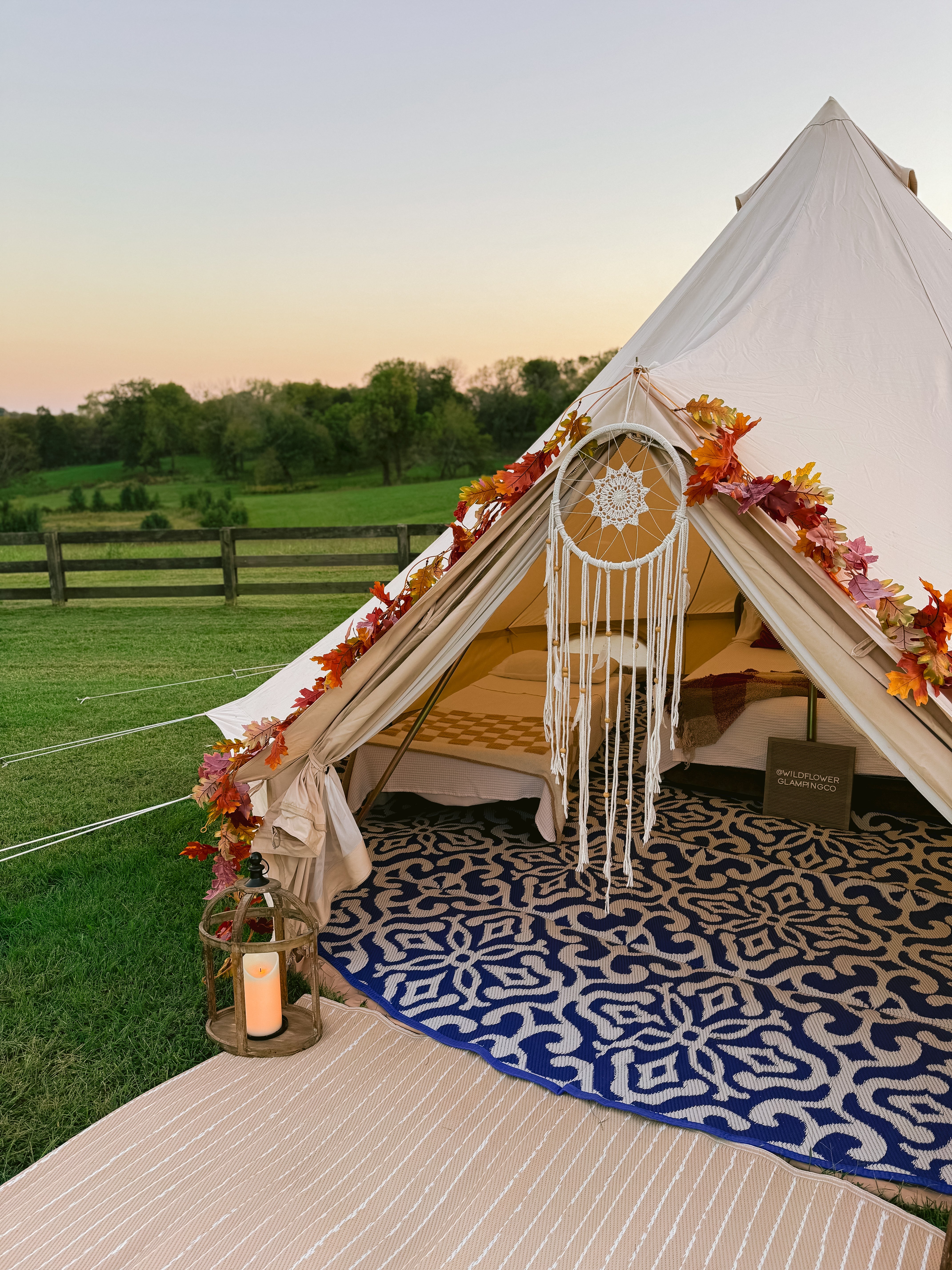 Decorated tent with fall leaves, a rug, and lantern on a grassy field with a wooden fence in the background.