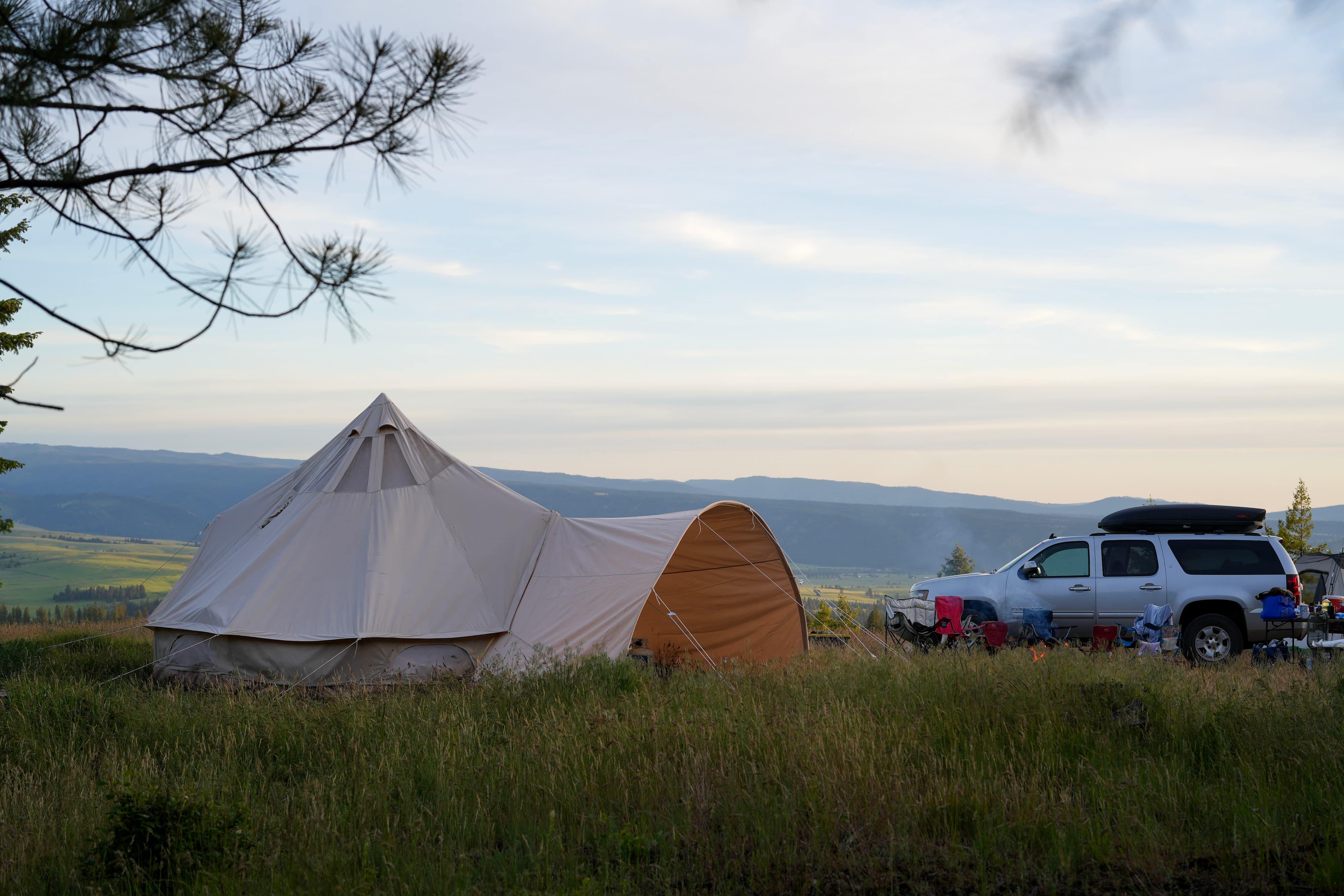 A large beige bell tent pitched in an open grassy field, with a white pickup truck parked nearby and a scenic mountain valley in the background under a cloudy sky.