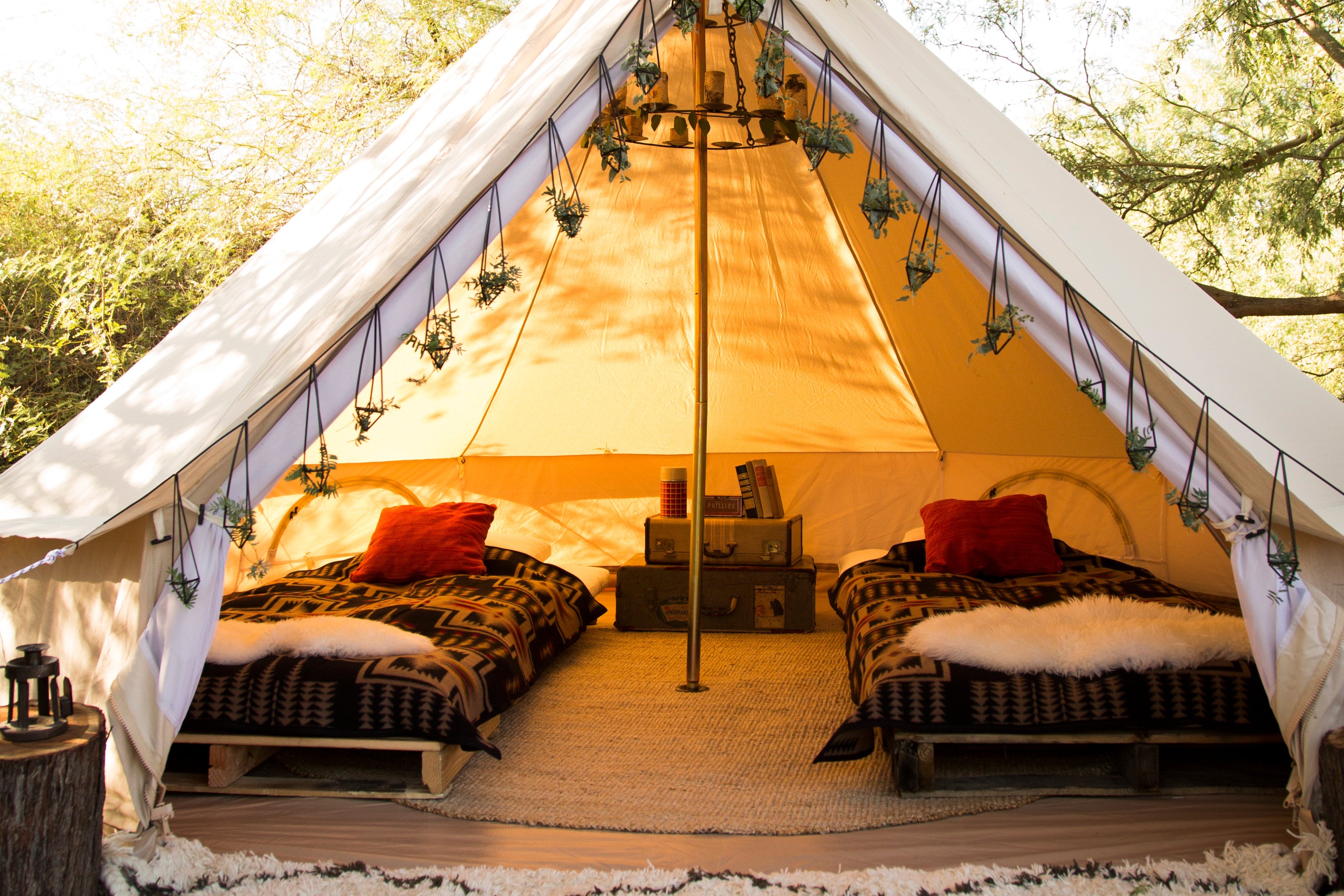 A large white canvas teepee tent set up outdoors under trees, with two beds inside, red pillows, string lights, and a patterned rug in front.