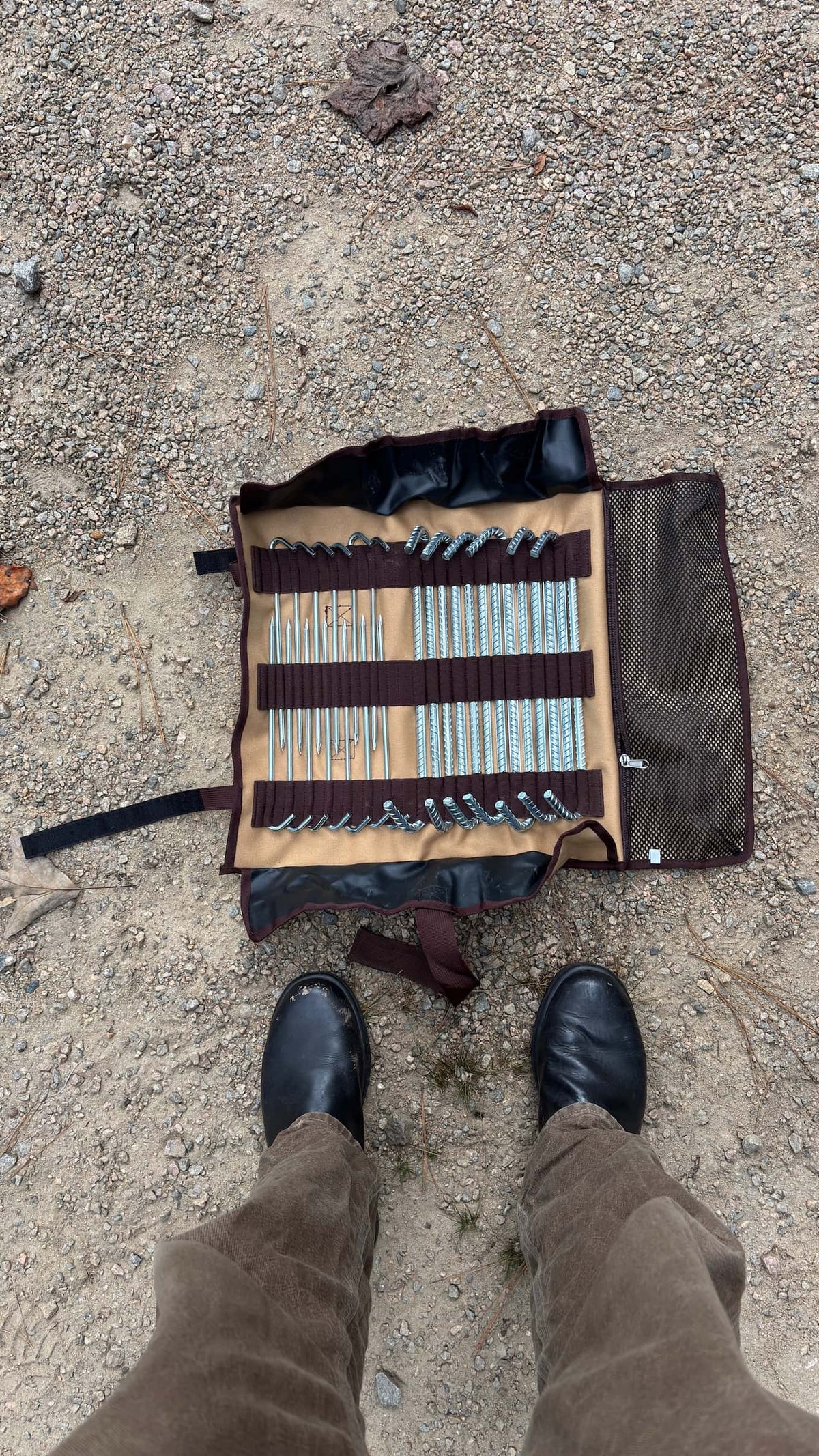 A set of metal tools organized in a brown and tan roll-up case, placed on sandy ground with a person's legs and black boots visible at the bottom of the frame.