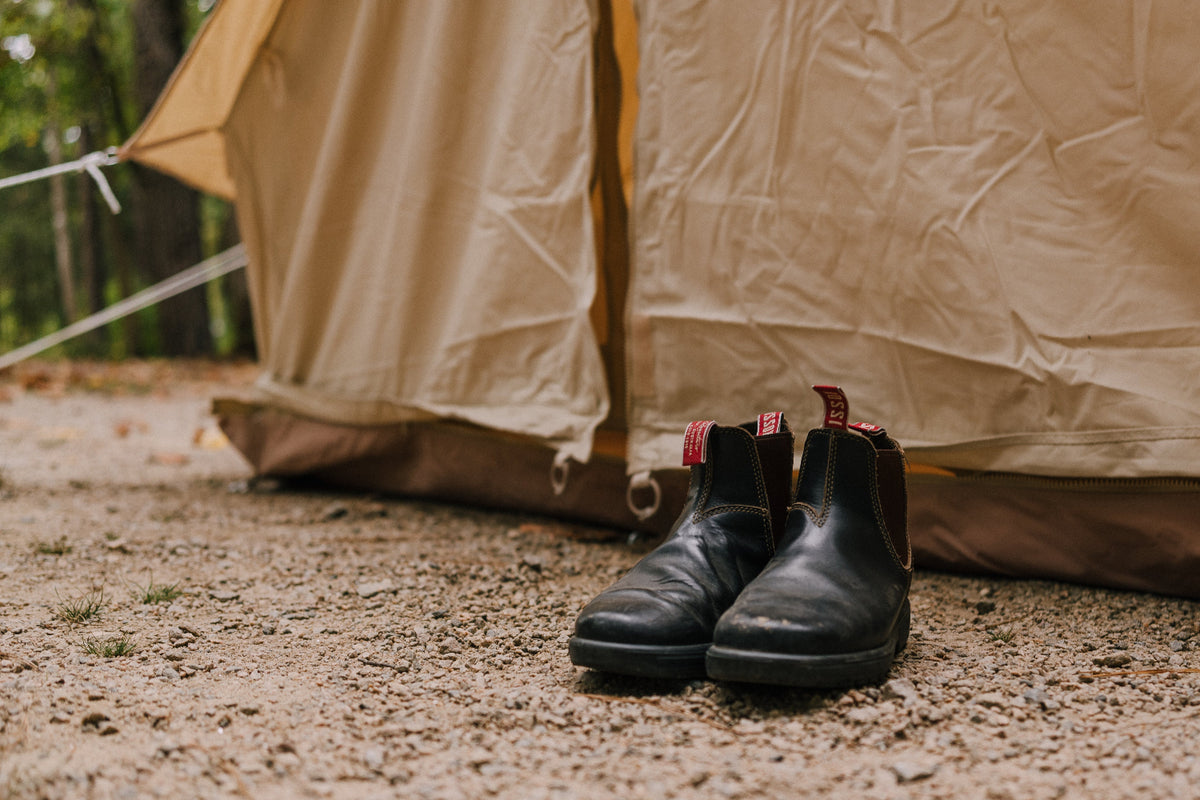 A pair of black leather Chelsea boots with red pull tabs sits on a gravel ground in front of a beige camping tent in a forest setting.