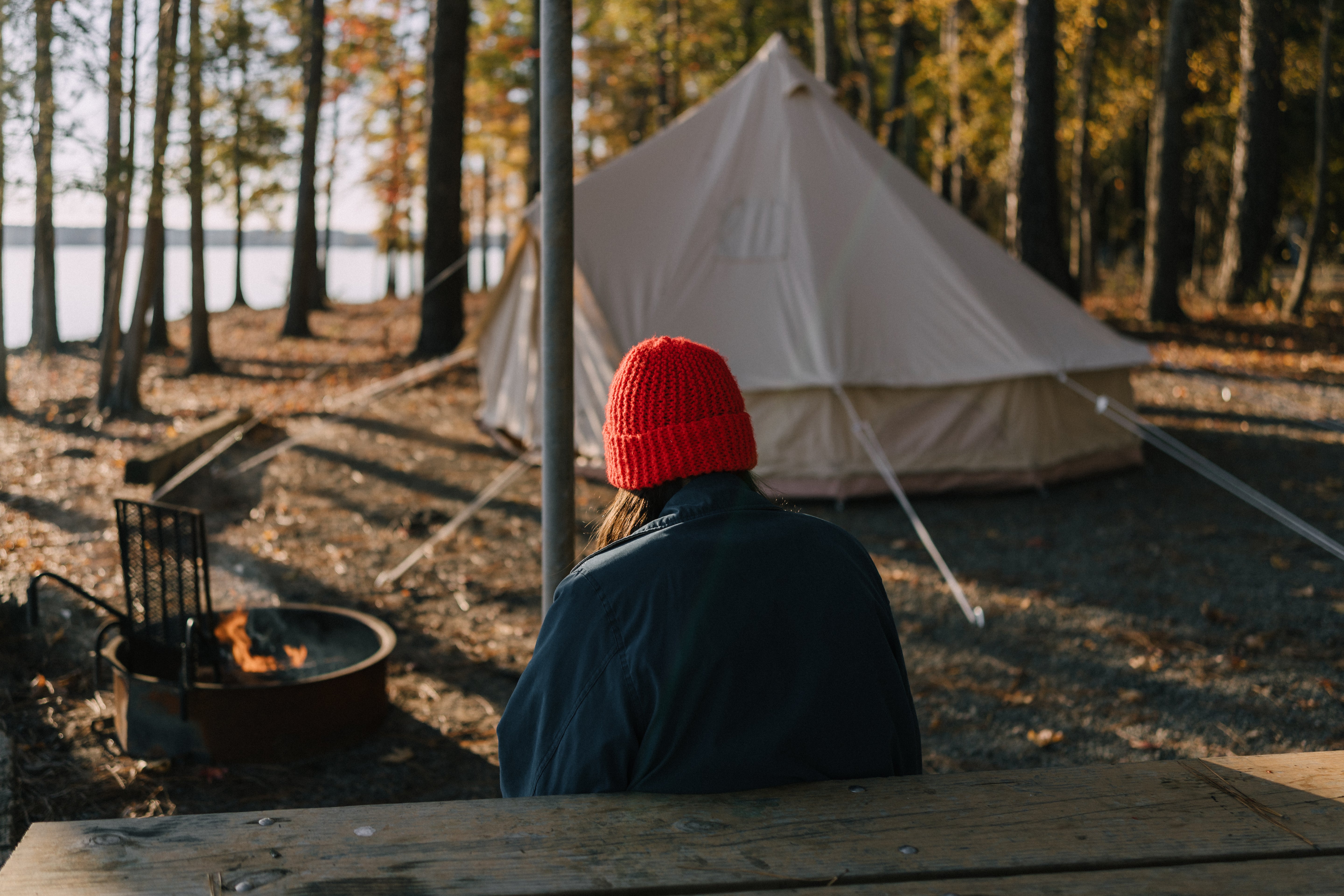 A person wearing a bright red knit beanie and a dark navy jacket sits at a wooden table in a campsite setting, facing a large beige bell tent. A fire pit with flames burns to the left, and a forest with autumn trees is visible in the background.