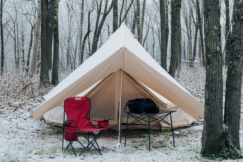 A large beige bell tent pitched in a wooded forest clearing, with a red camping chair and a small folding table set outside the tent entrance.