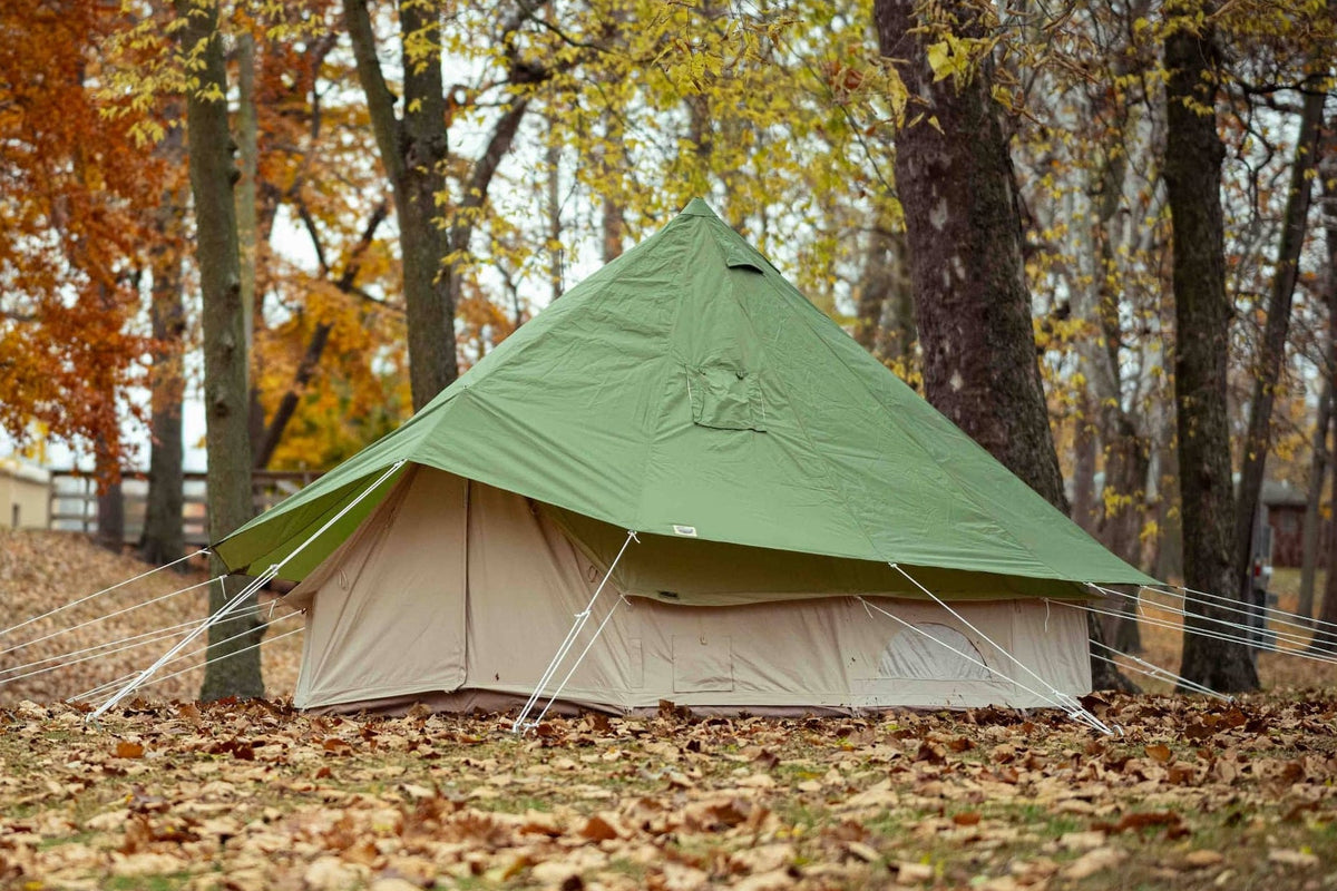 A green and beige bell tent pitched outdoors in a wooded area covered with fallen autumn leaves.