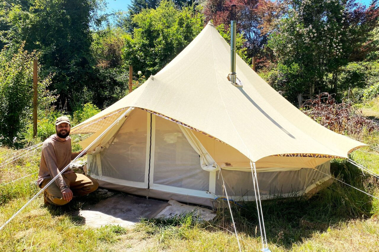 Man sitting in front of a large beige tent in a natural setting with trees and grass.