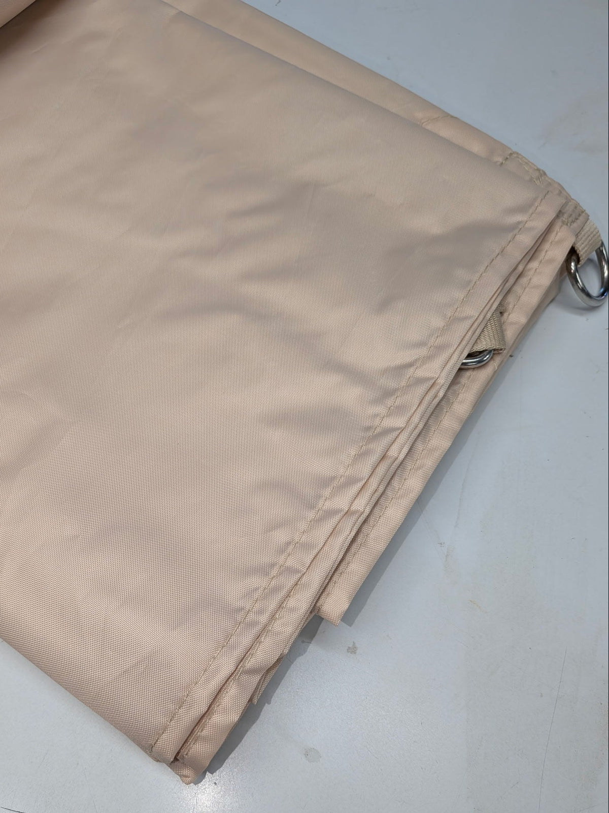 A folded beige fabric panel or curtain with visible stitching and a small metal grommet, placed on a light gray surface.