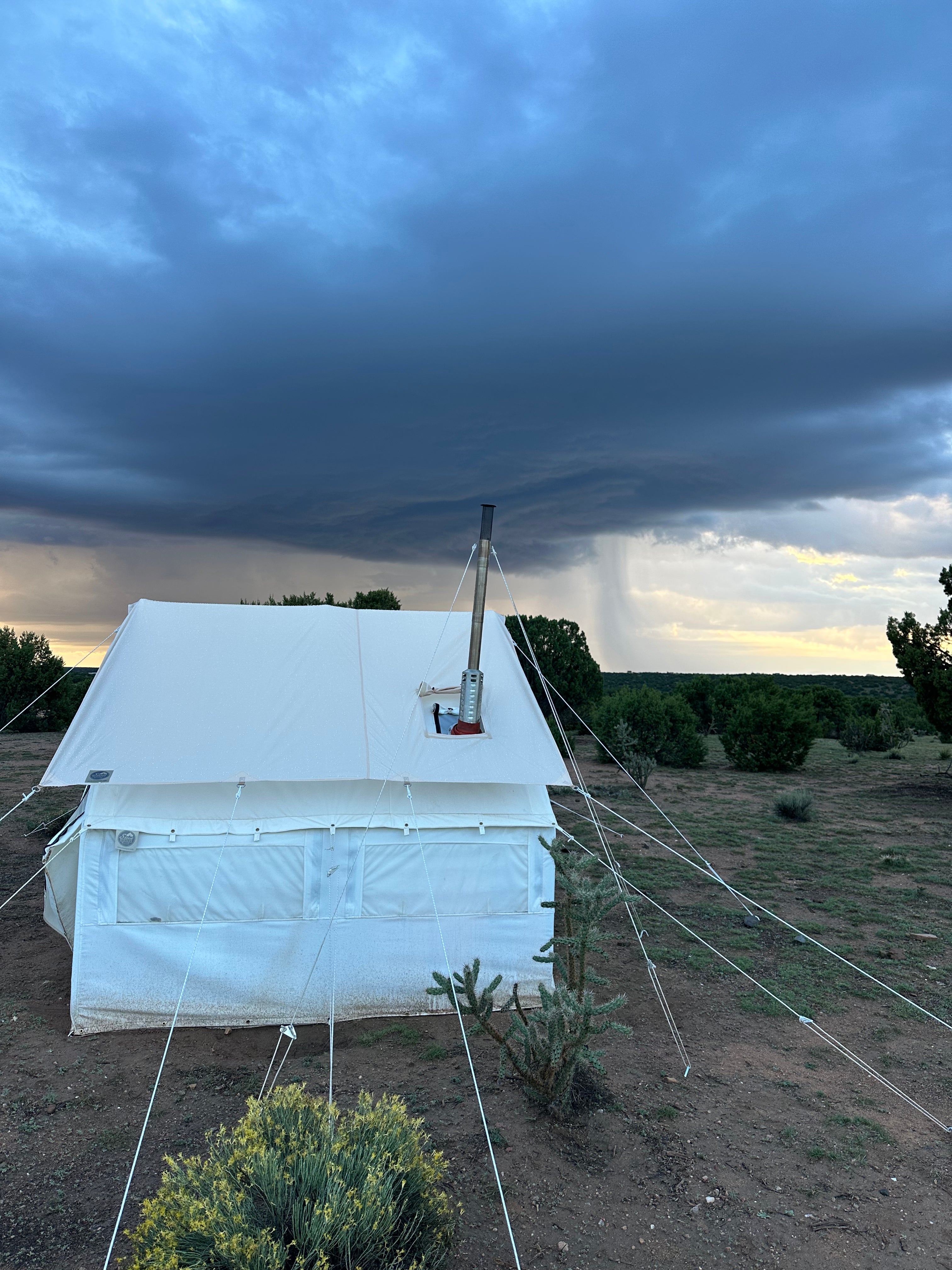 A large white canvas glamping tent pitched outdoors in a dry grassy landscape under a dramatic dark storm sky with rain visible in the distance.