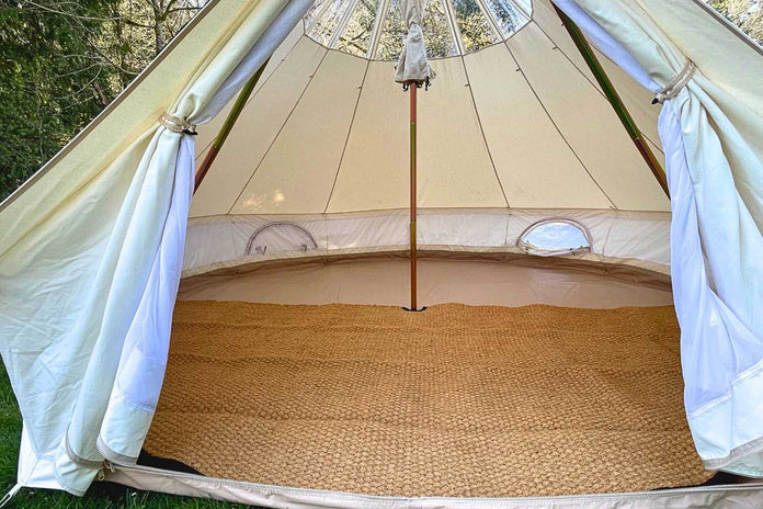 Inside view of a beige bell tent with a woven mat on the floor, surrounded by greenery.