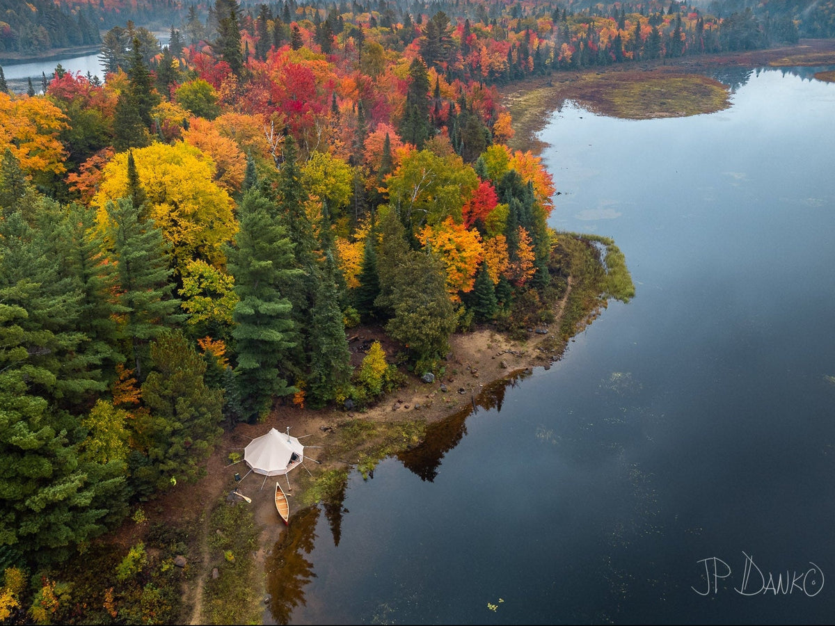 Aerial view of a calm lake bordered by dense autumn forest in vivid yellow, orange, red, and green tones, with a small white tent and canoe near the shoreline under an overcast sky.