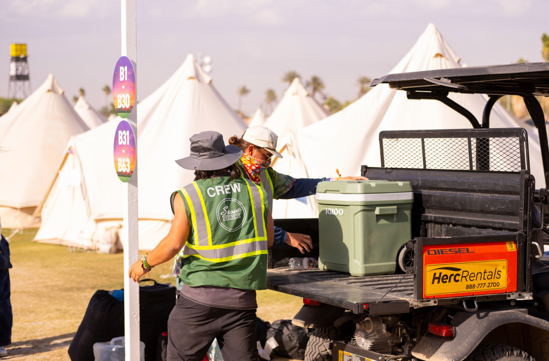 Two event crew members loading a cooler into a truck with tents and a clear sky in the background.
