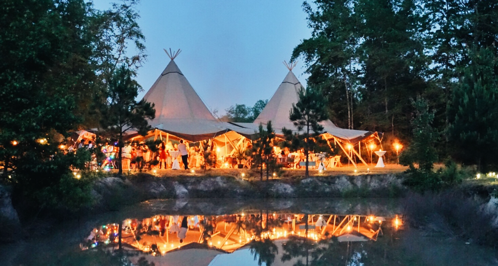 Tipi-style tents at night with warm lighting reflected in a pond, surrounded by trees.